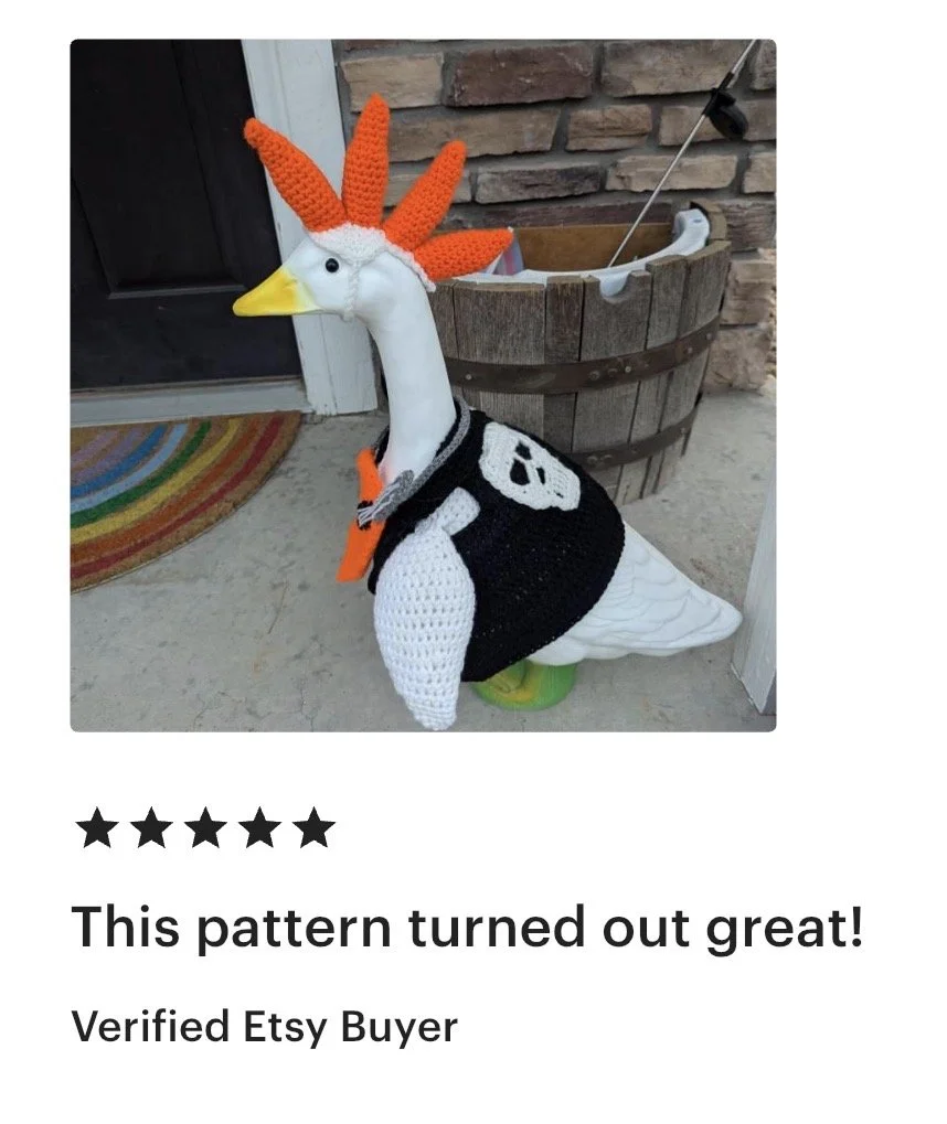 Stuffed goose dressed in a pirate costume with a crocheted skull and crossbones vest, a hat, and an orange beard, standing on a porch next to a brick wall and a wooden barrel planter.