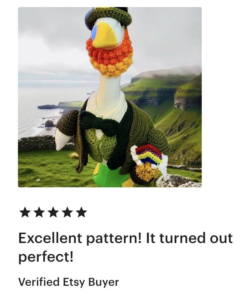A plush goose wearing a crocheted scarf, hat, and sweater, holding a rainbow-colored crocheted item, standing outdoors with coastal cliffs in the background.