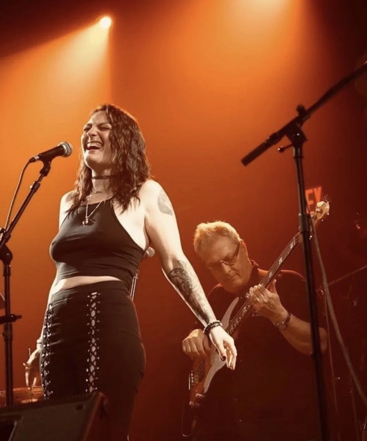 A woman singing passionately into a microphone on stage, with a guitarist playing in the background, under warm orange stage lighting.