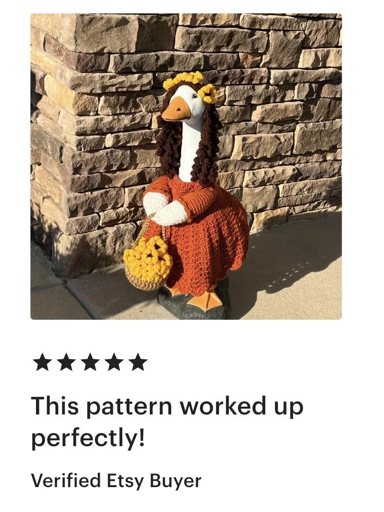 A decorative stuffed goose dressed in a red crochet dress and a yellow flower crown, holding a yellow flower purse, standing against a stone wall.