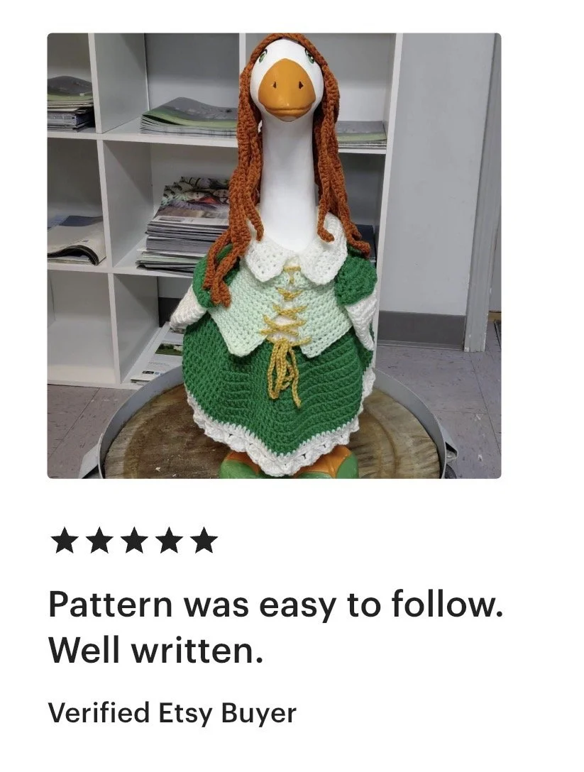 A stuffed duck dressed in a crocheted princess costume with a green skirt and white top, standing on a wooden surface against a background of white shelves filled with papers and magazines.