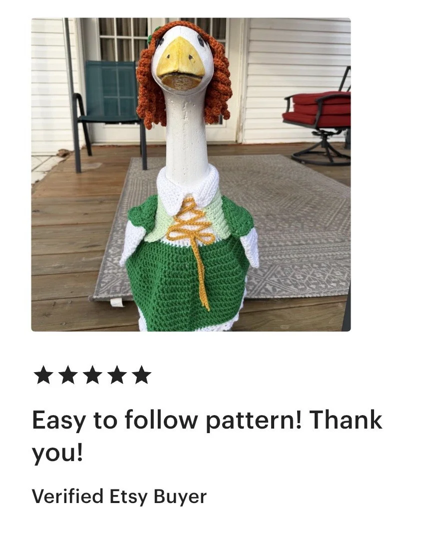 A decorative goose statue dressed in a green and white knitted sweater, with an orange and white striped tie, and a sock hat. The goose has curly orange yarn hair and a painted face with a yellow beak.