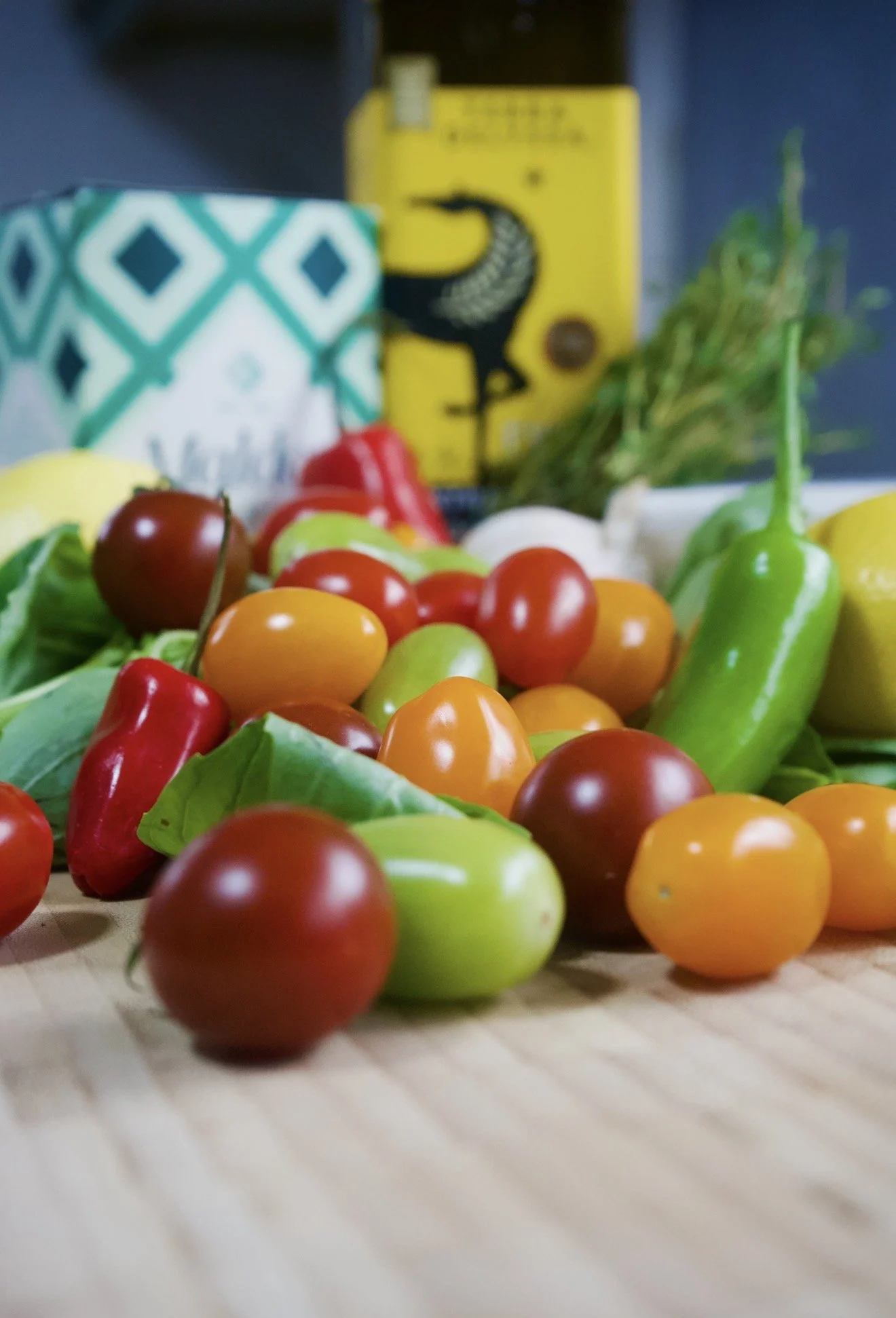 Close-up of a variety of colorful cherry tomatoes on a wooden surface, with a blurred background including a patterned cup, a yellow box, and some green herbs.