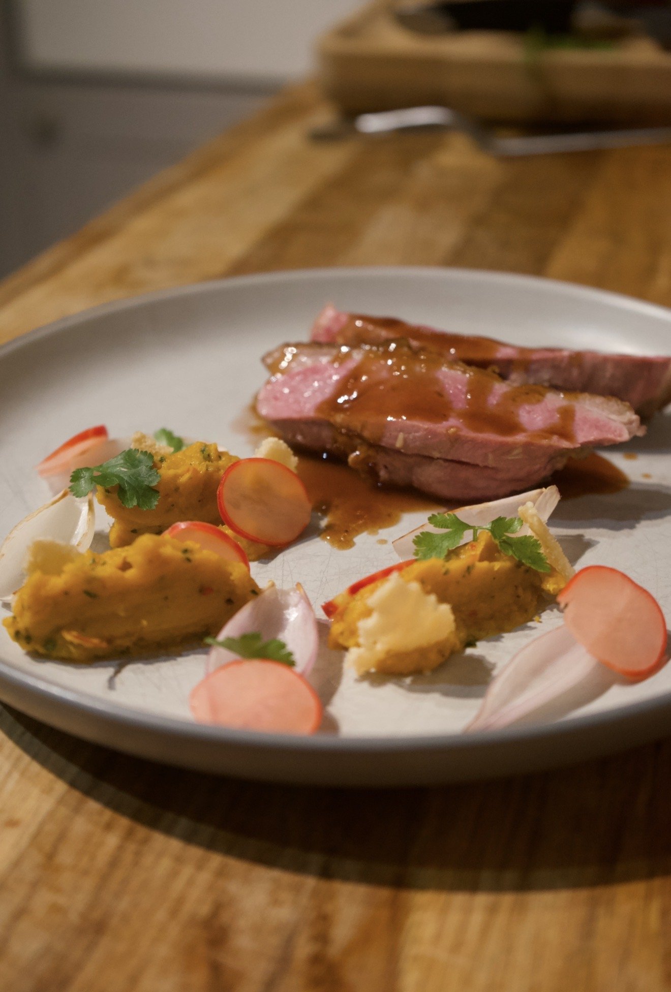 A white plate with sliced duck topped with red wine jus, accompanied by roasted vegetables and garnished with parsley and radish slices, on a wooden table.