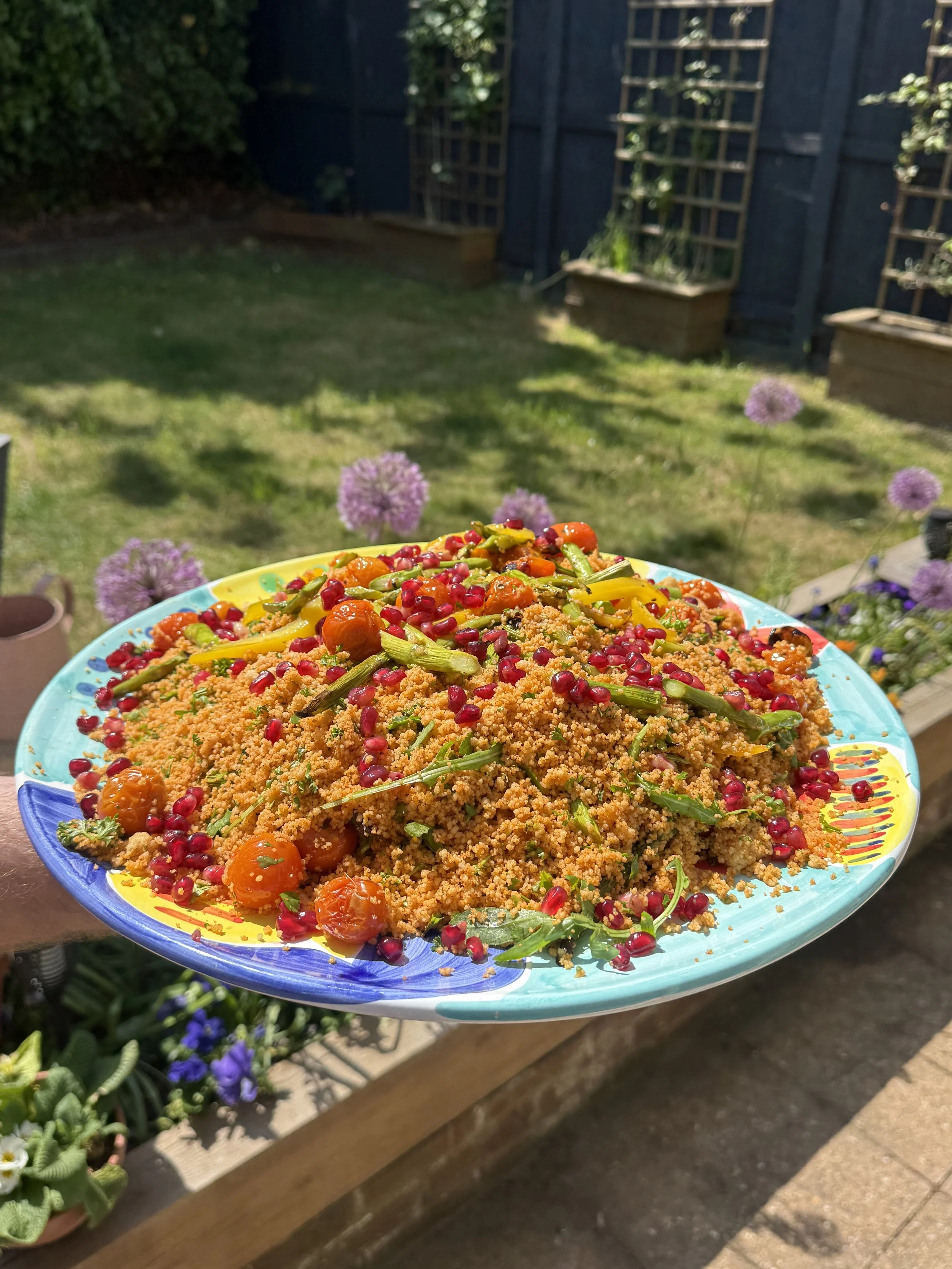 A colorful plate of couscous salad topped with cherry tomatoes, pomegranate seeds, and green herbs. The background shows a garden with green grass, purple flowers, and wooden planters, all under bright sunlight.