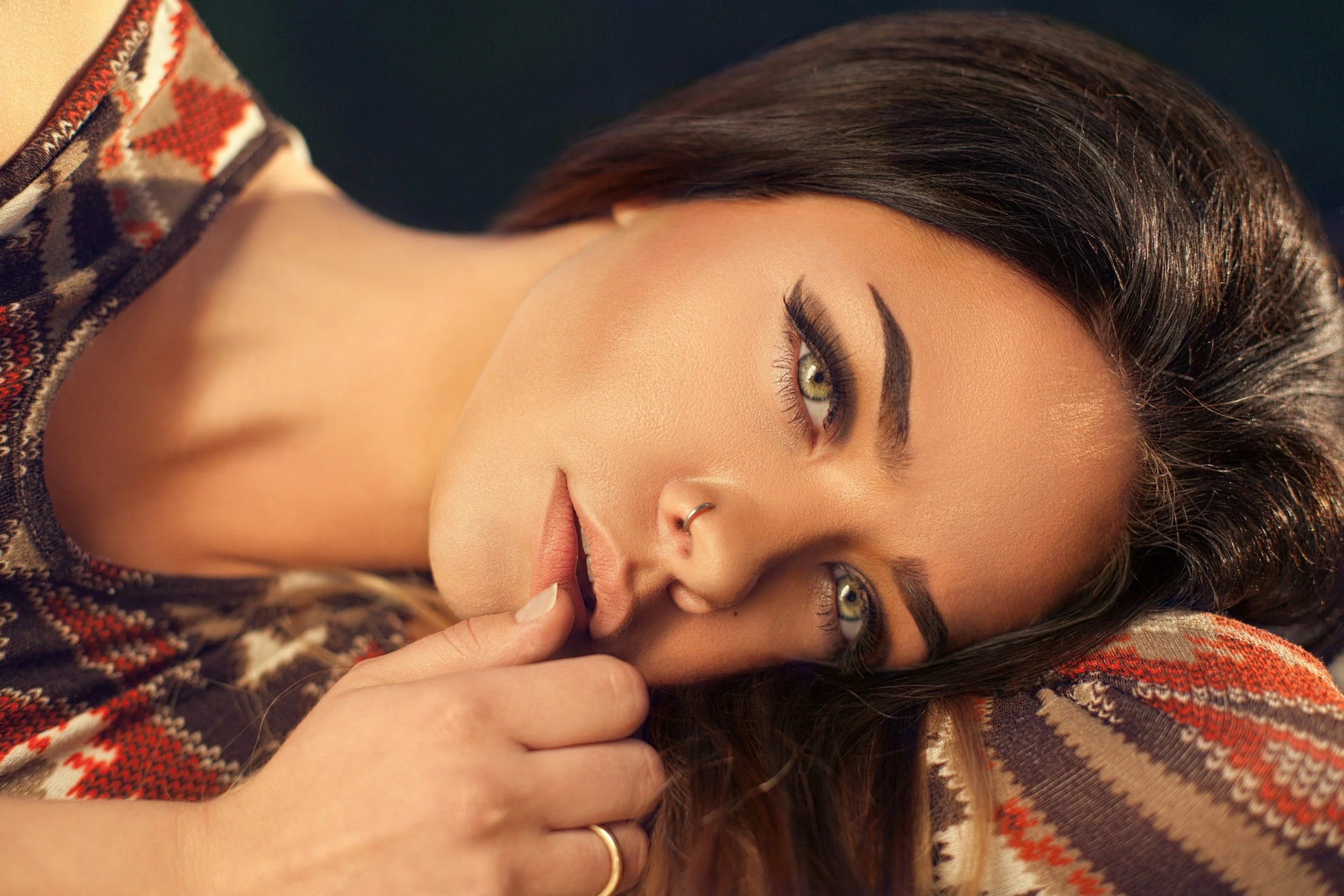 Close-up of a woman with dark hair and light skin, lying down, touching her lips with her index finger, wearing a patterned bohemian sweater.