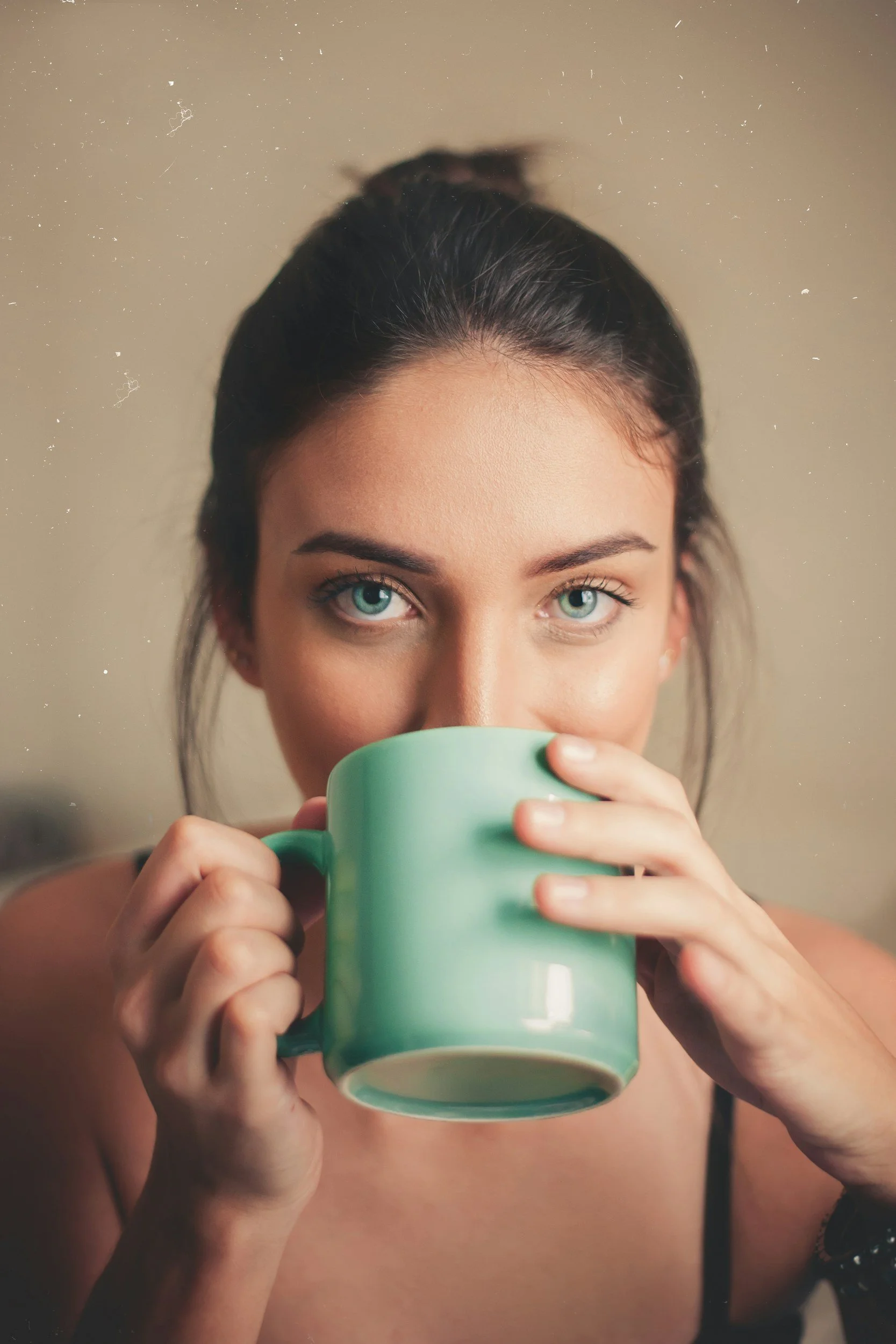A young woman with blue eyes and dark hair tied back, holding a green mug close to her face while sipping from it, with minimal clothing visible.
