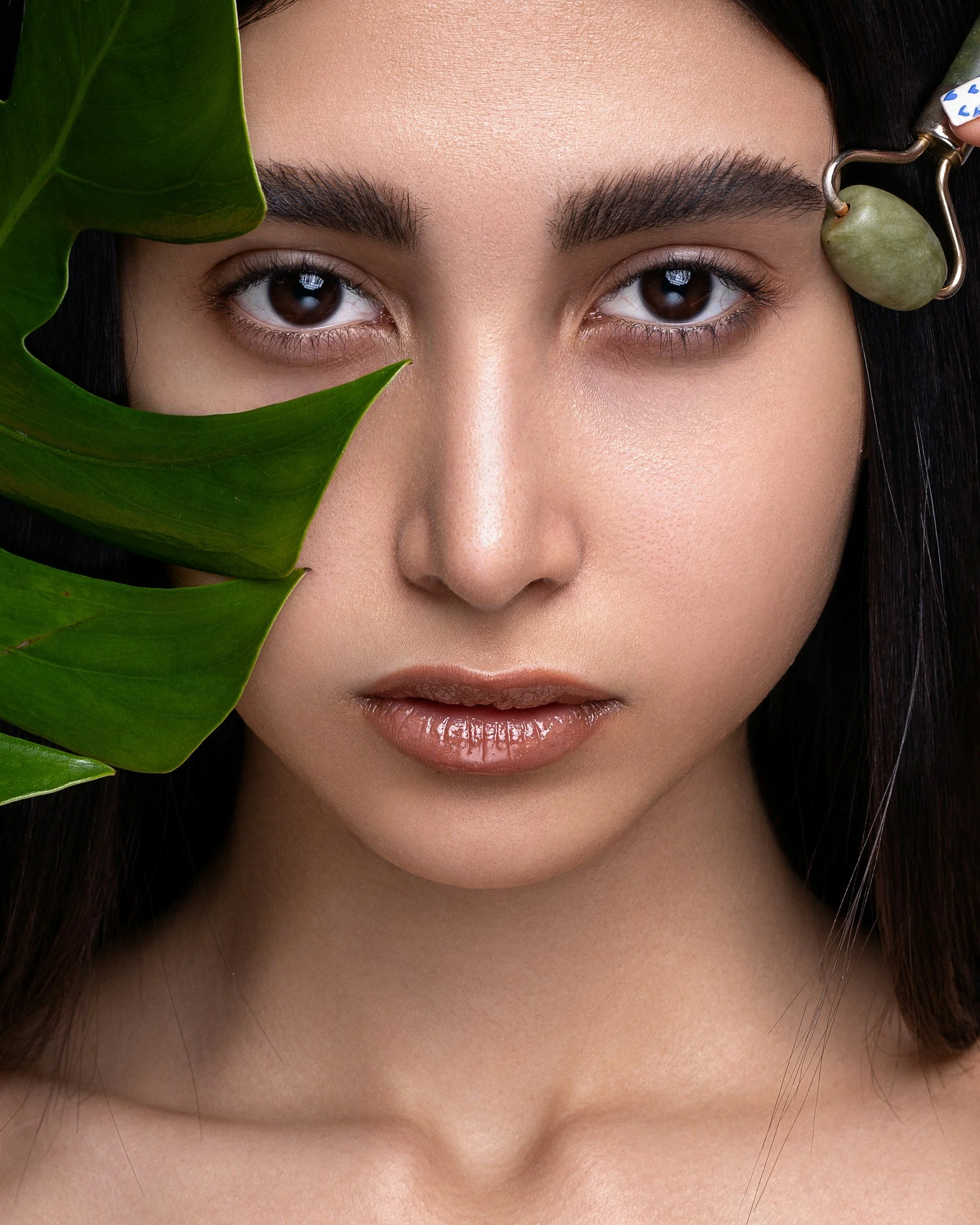 Close-up of a woman's face with dark hair, showing her eyes, eyebrows, and lips, partially obscured by large green leaves and a jade roller on her right temple.