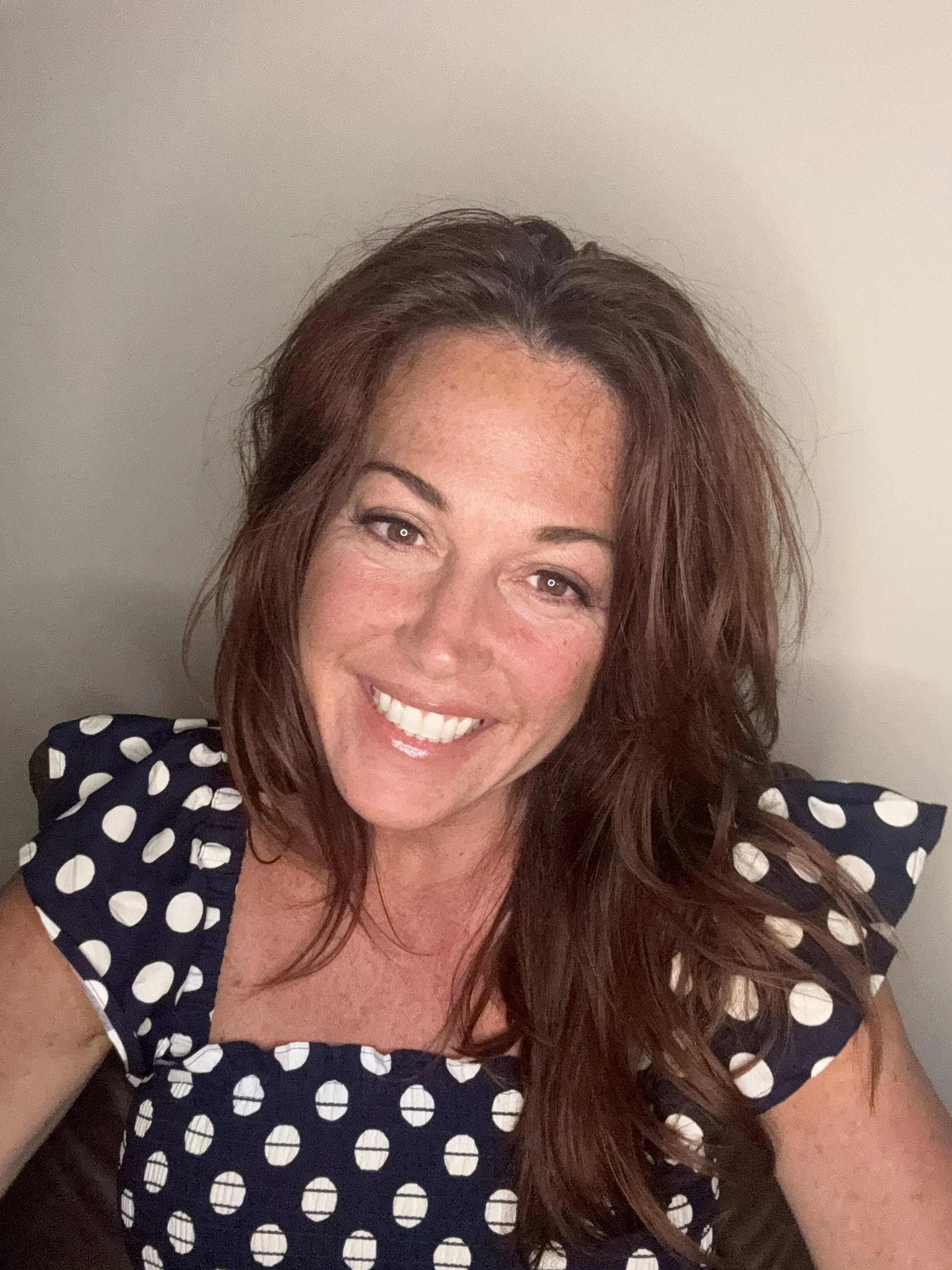 A woman with long, wavy, reddish-brown hair smiling at the camera, wearing a navy blue dress with white polka dots, sitting on a dark brown leather chair against a plain beige wall.