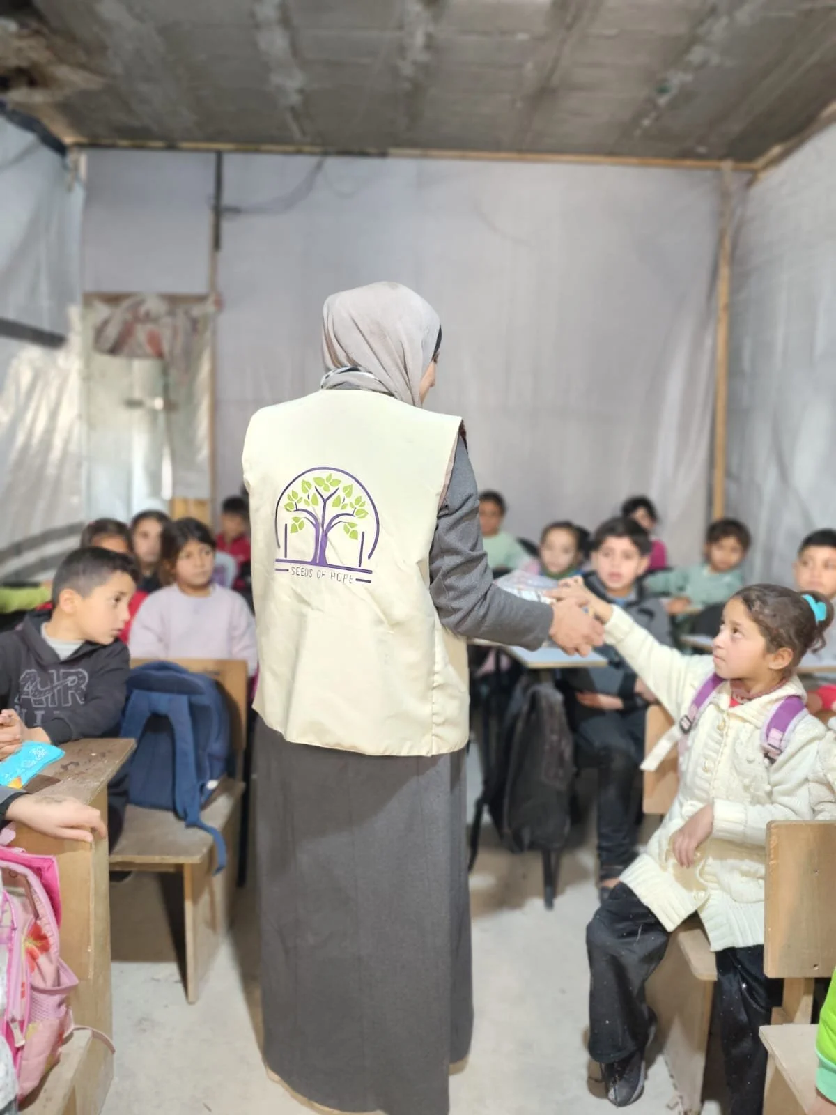 A woman wearing a vest with a logo handing out items to students in a classroom with a makeshift ceiling and plastic walls.