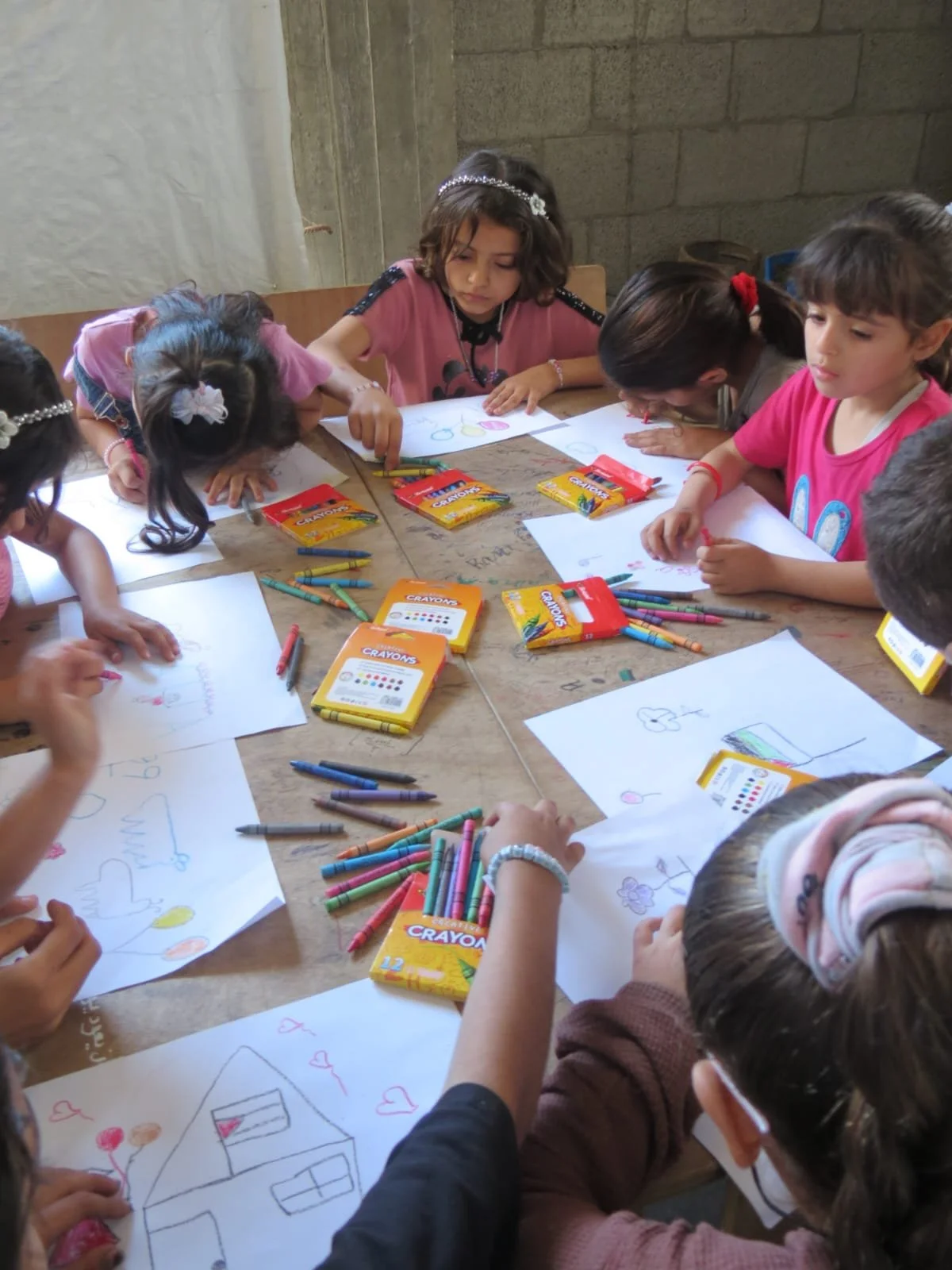 Children drawing and coloring pictures at a wooden table with crayons and drawings of houses, balloons, and animals.