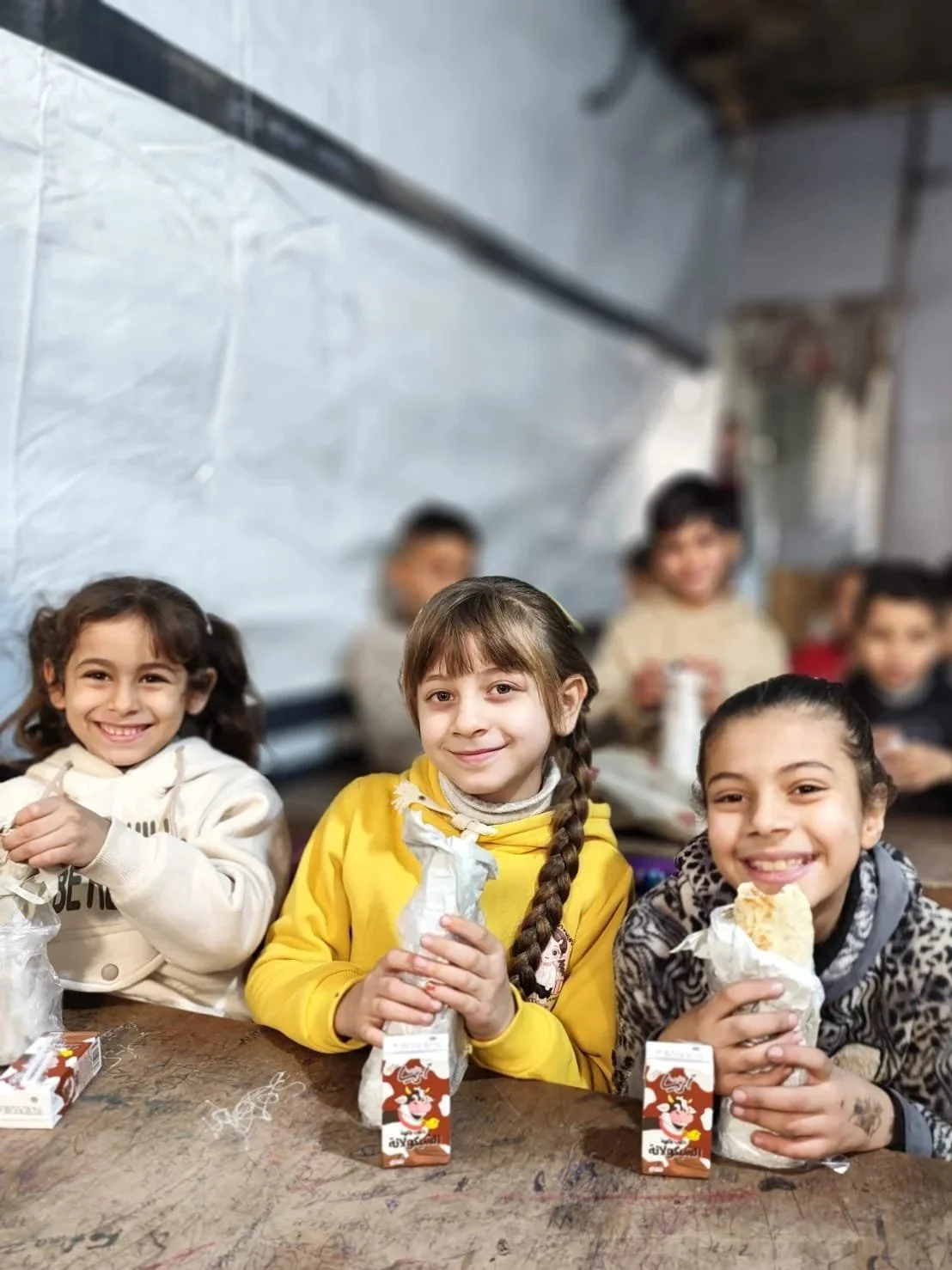 Three young girls sitting at a table, smiling and holding wrapped sandwiches and juice boxes, with other children in the background.