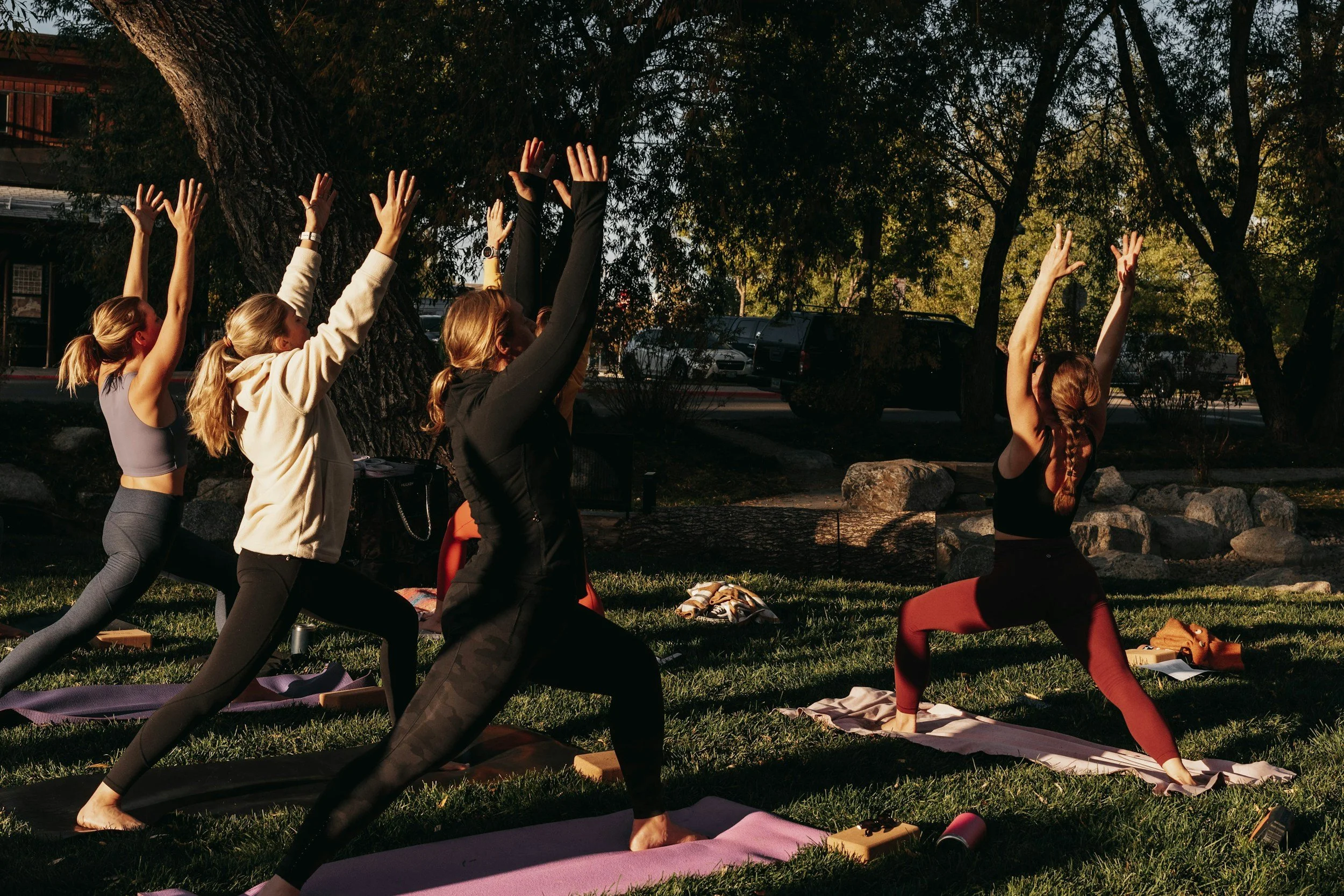 Group of women practicing yoga outdoors at sunset in a park, sitting on yoga mats with arms raised.