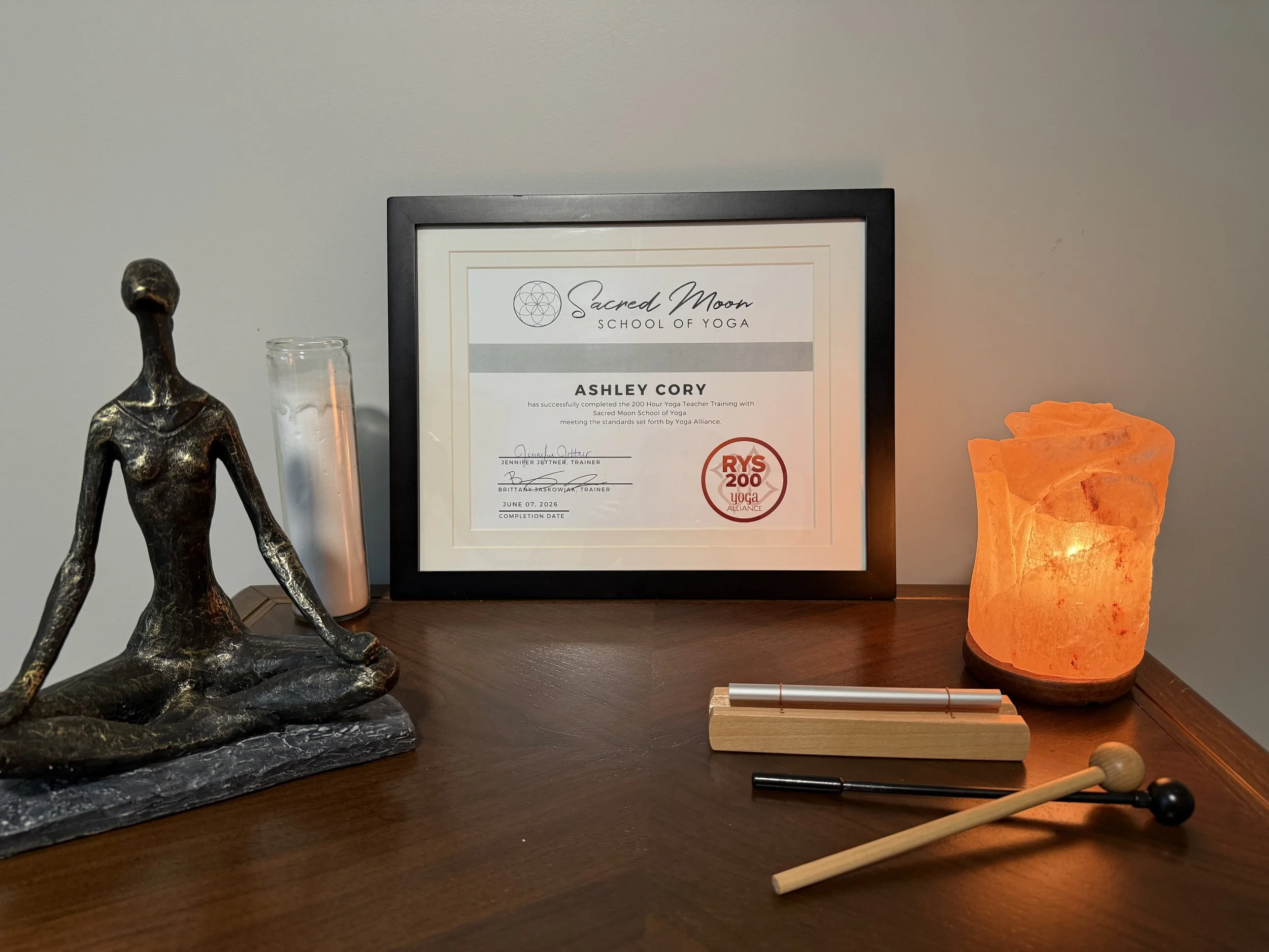 A diploma from Sacred Moon School of Yoga awarded to Ashley Cory, featuring a black frame, a decorative logo, and a red seal. The desk displays a bronze statue of a woman, an orange Himalayan salt lamp, and various wooden and rubber massage tools.