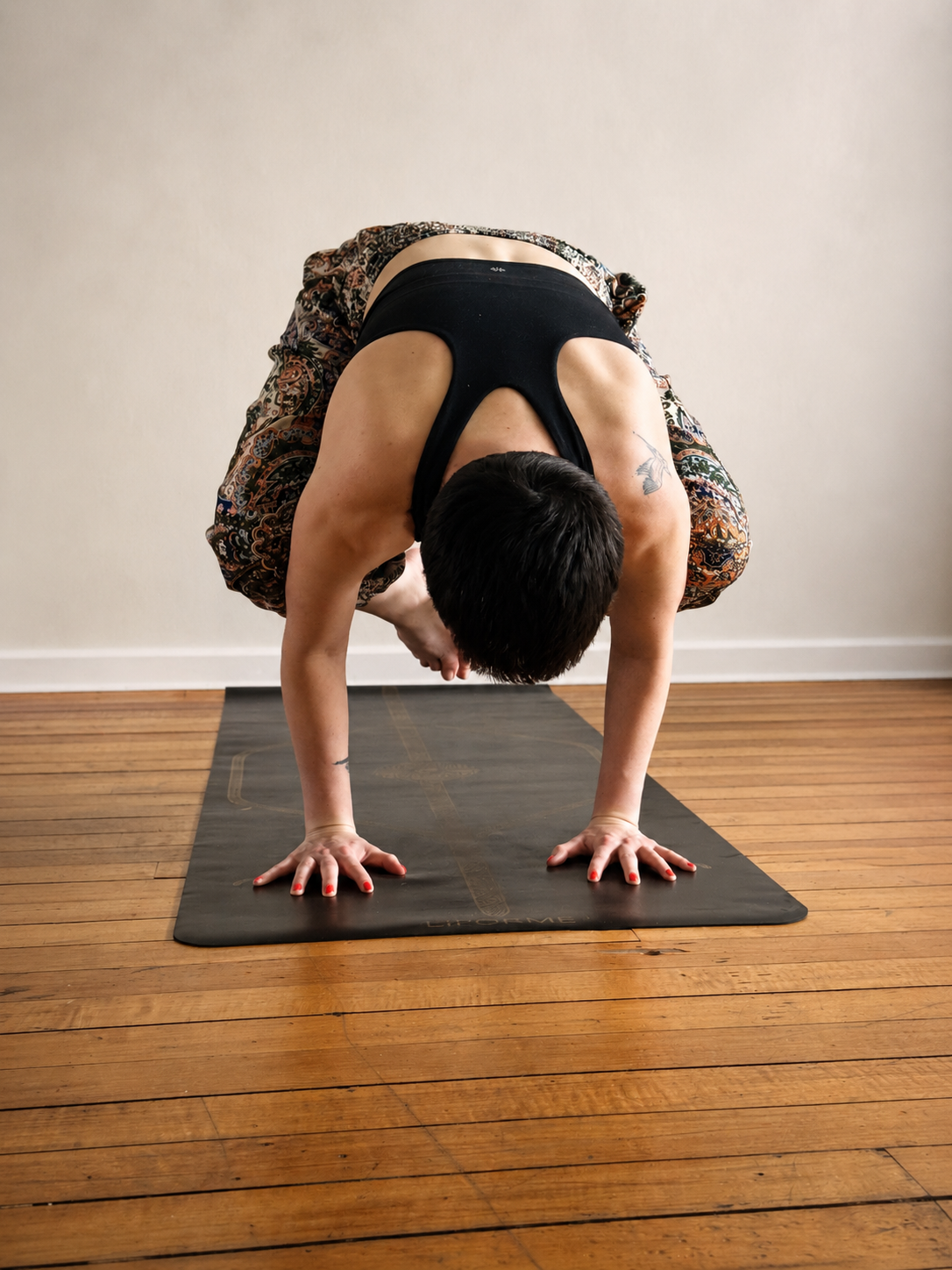 Person practicing yoga in crow pose on a black yoga mat in a room with wooden floors and a plain white wall.