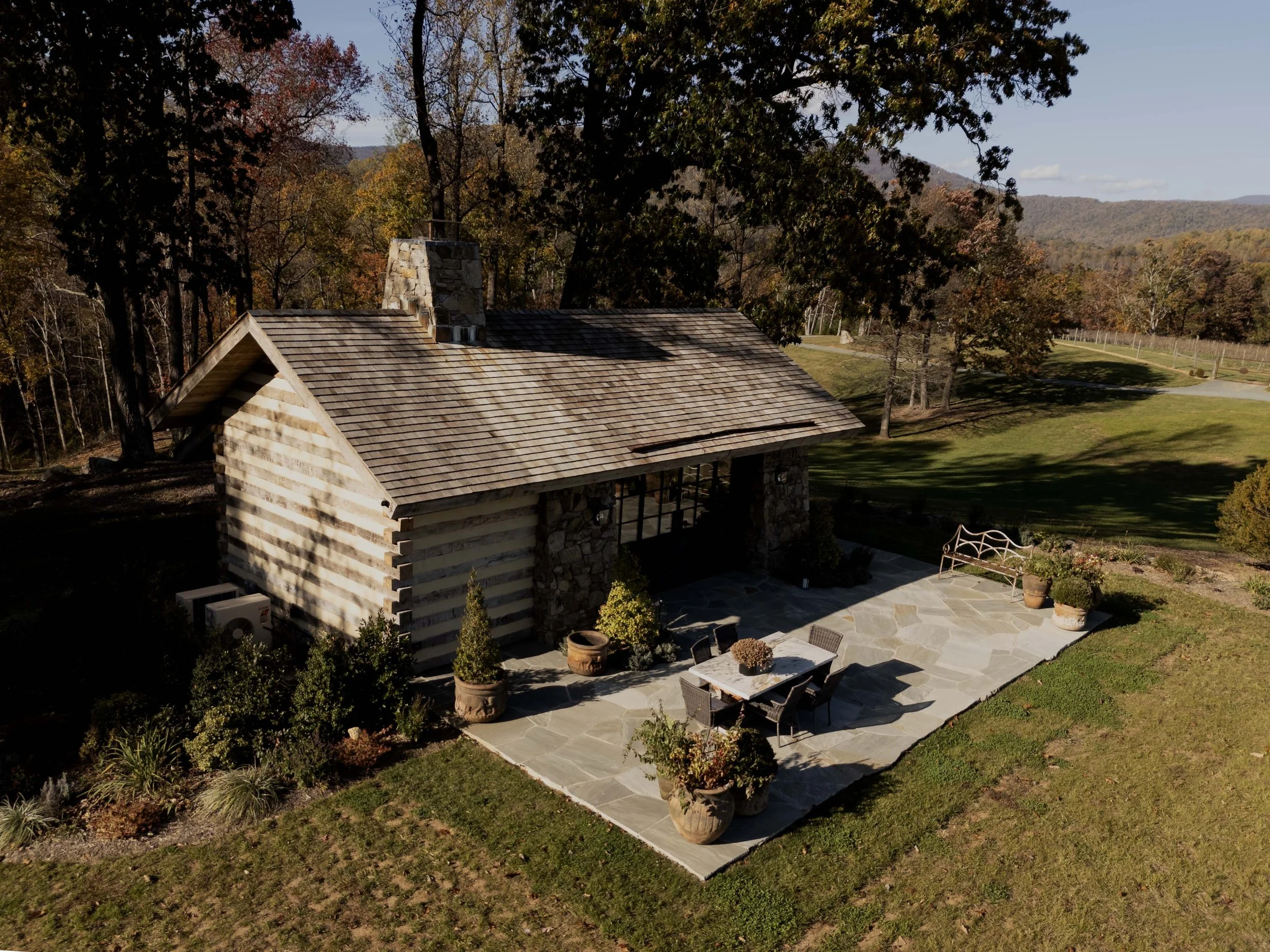 Aerial view of a house with stone and wooden exterior, surrounded by a garden with potted plants, trees, and a stone patio, with hills and trees in the background.