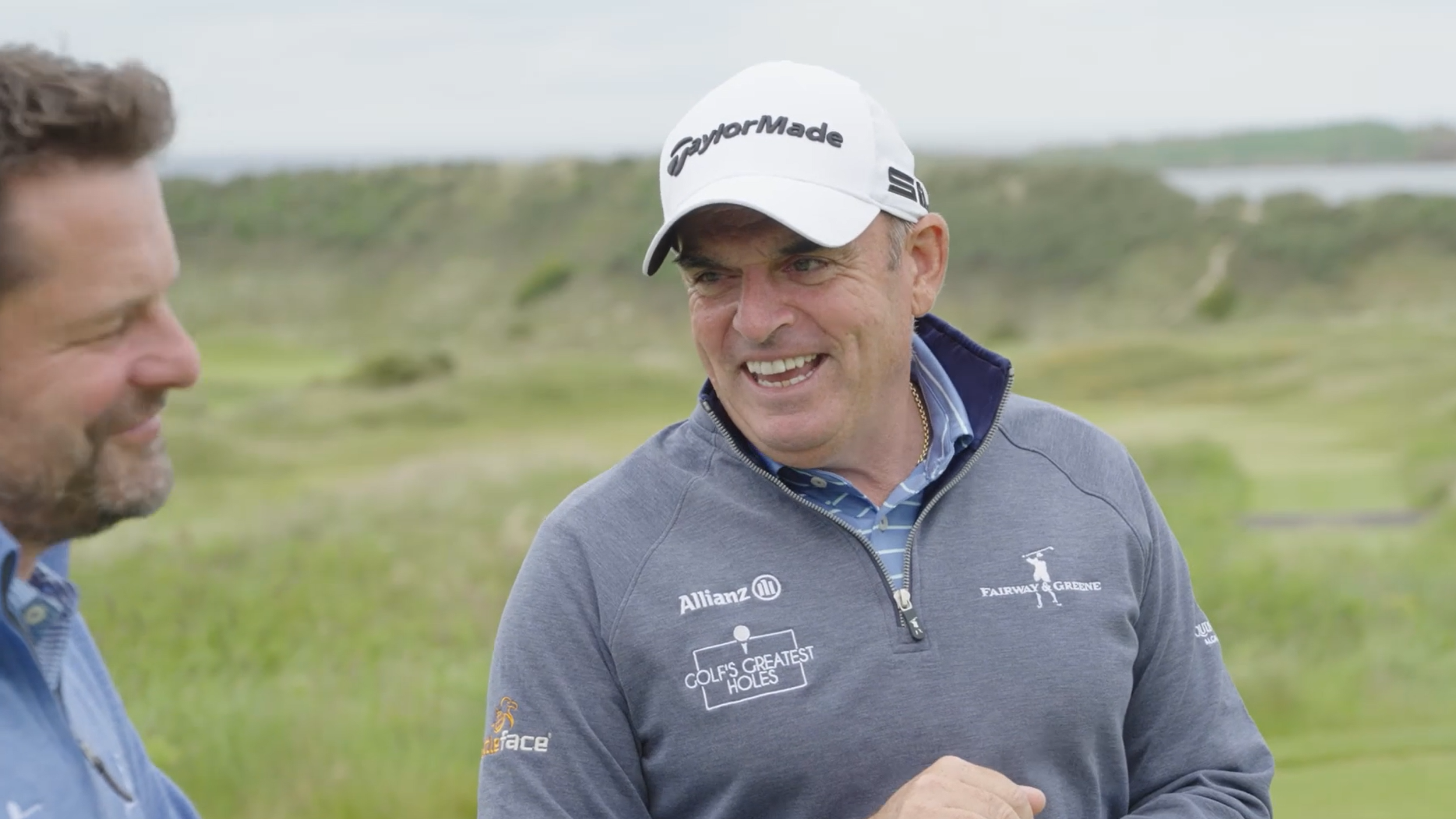 Two men outdoors, one in a golf hat and gray jacket with sponsor logos, smiling and engaging in conversation, with a green landscape and cloudy sky in the background.
