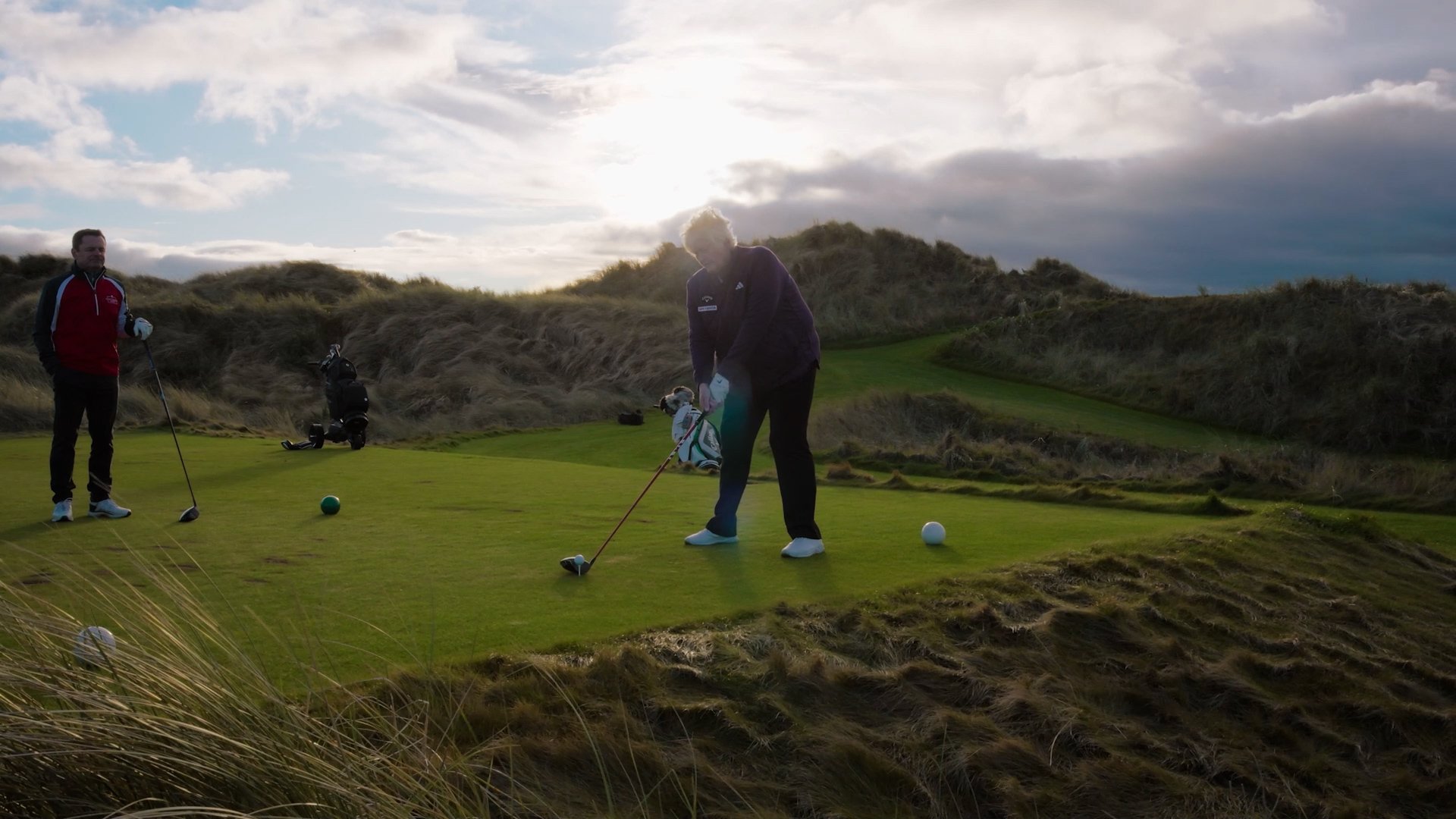 Two men playing golf on a green course with grassy hills and a cloudy sky in the background.
