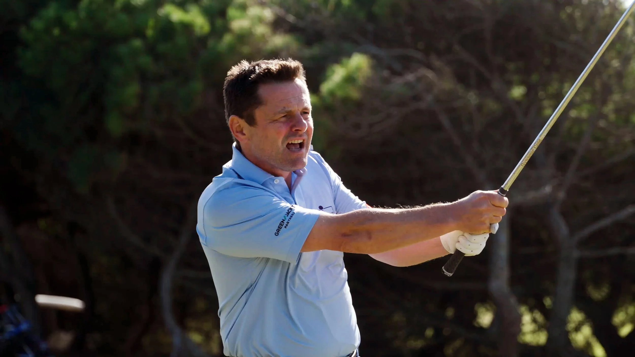 Man in a light blue golf shirt swinging a golf club outside, with trees in the background.