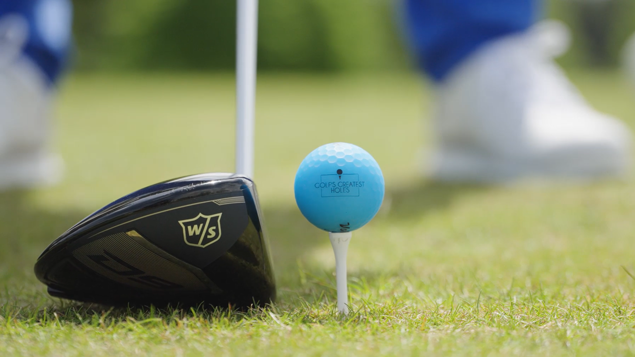 Close-up of a golf club lying on the grass with a golf tee and a Hello, World golf ball labeled 'Golf's Greatest Holes' in the background, on a golf course.