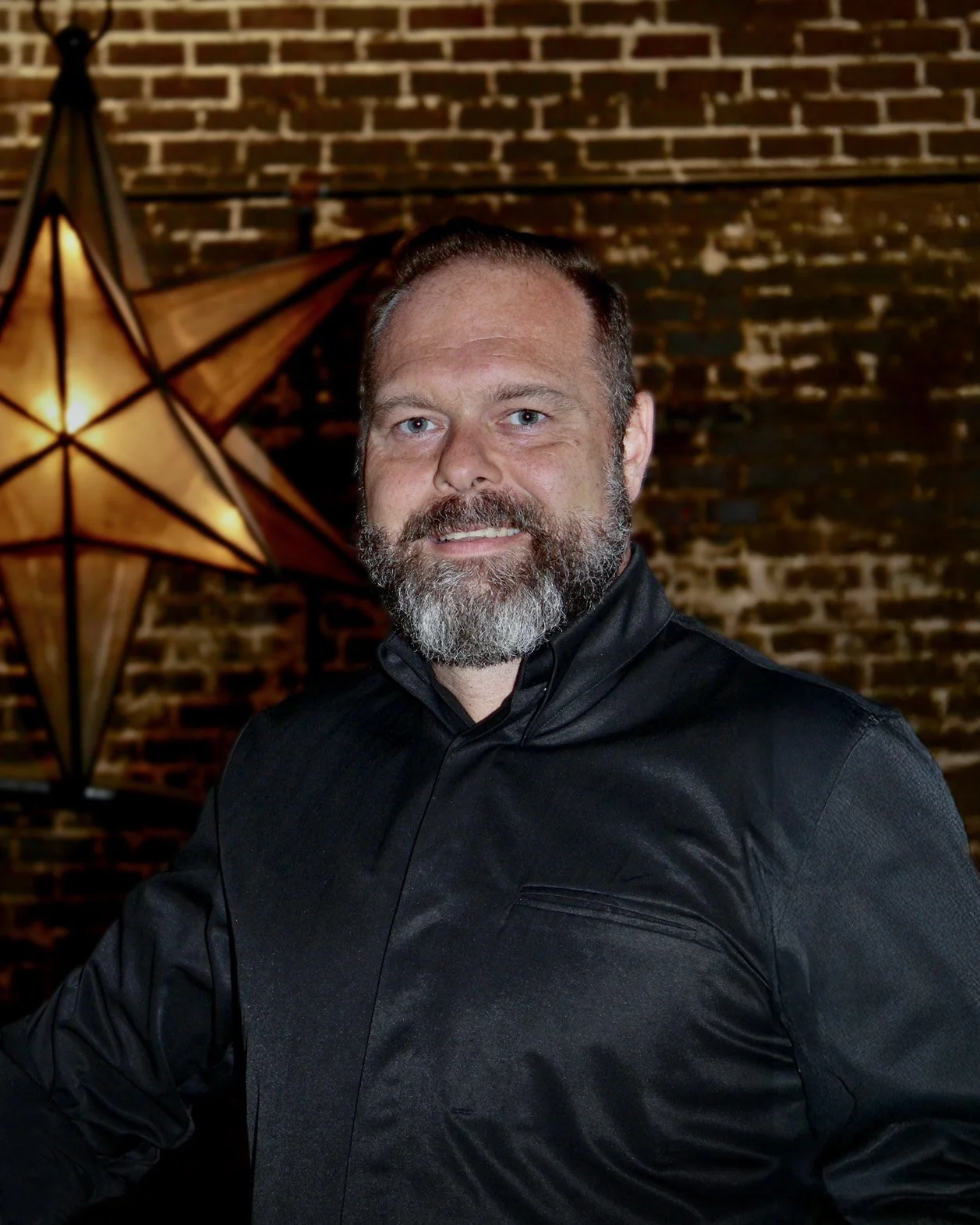 A man with a beard and mustache, wearing a black shirt, stands in front of a brick wall with a decorative star-shaped lamp in the background.