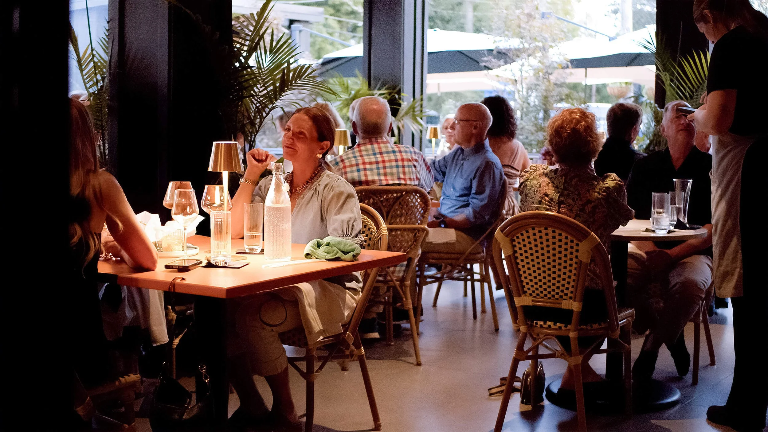 Indoor dining scene with multiple people seated at tables, some engaged in conversation, and a waitress serving. Large windows with natural light and green plants visible outside.