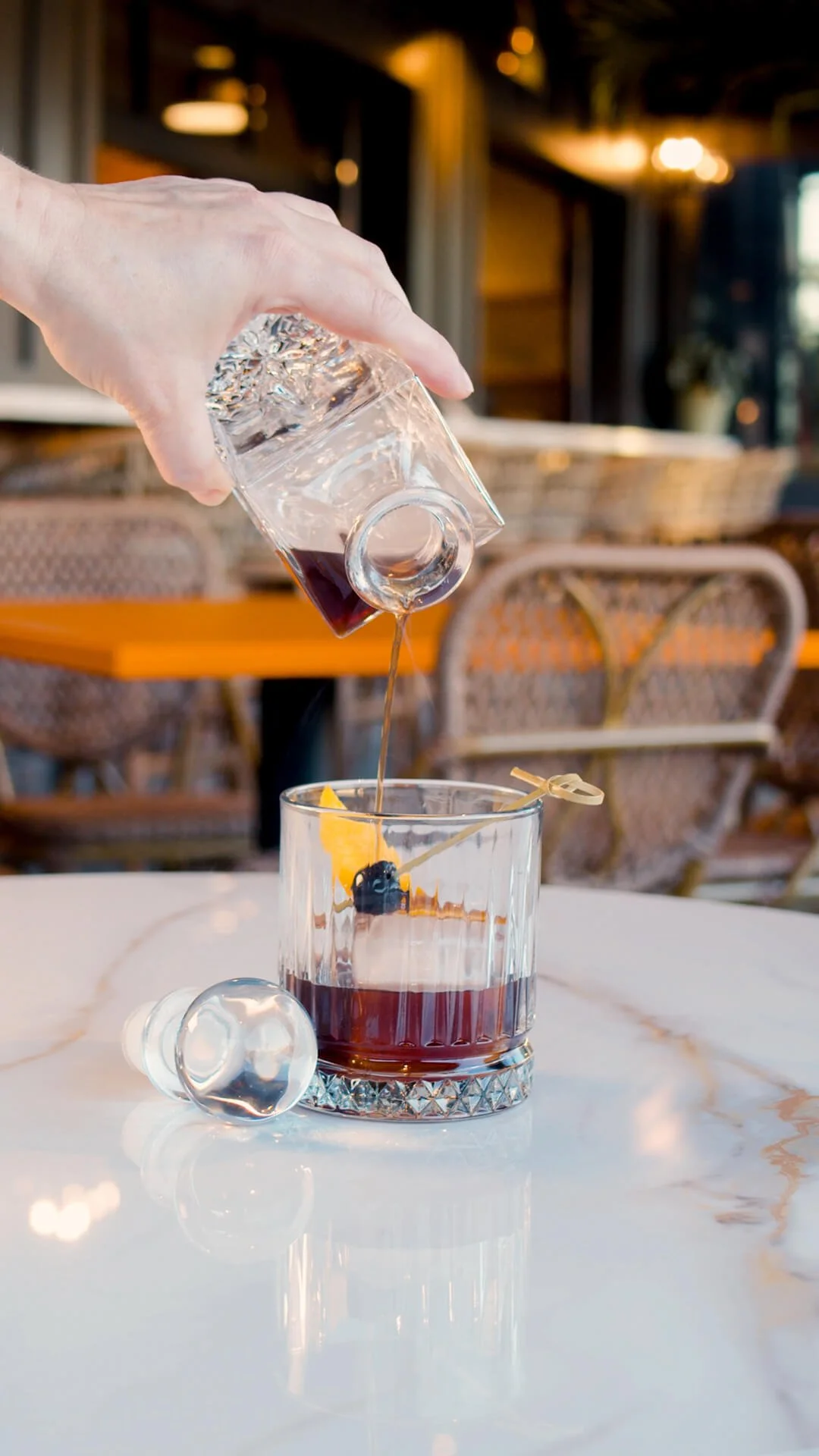 A hand pouring red wine into a glass, with a cherry and orange slice garnish on a marble table, in a cozy restaurant setting.