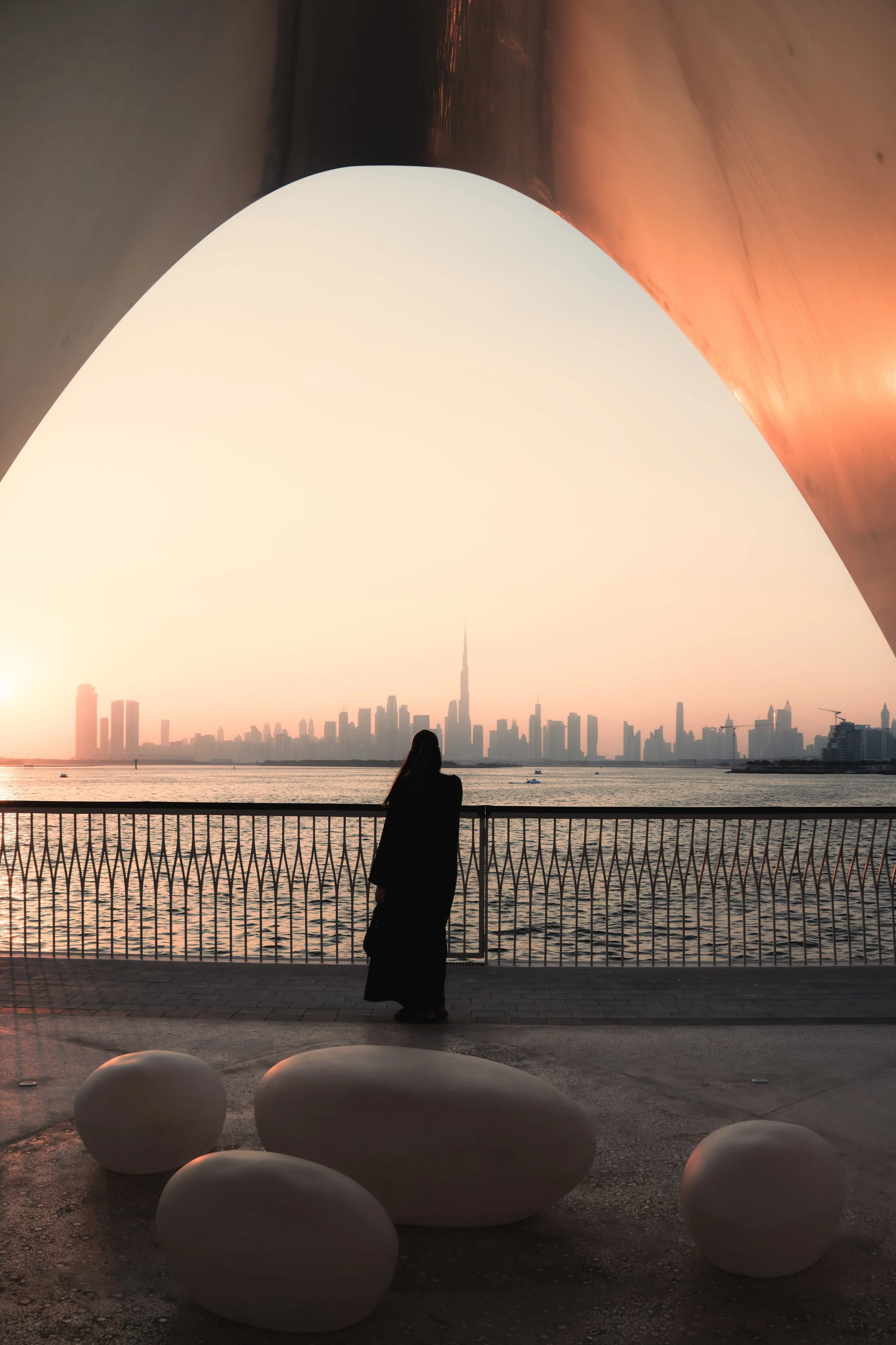 A woman stands by a waterfront railing at sunset, with a city skyline and the One World Trade Center in view in the background. Large white stone sculptures are in the foreground, and the sky transitions from warm orange to pale blue.