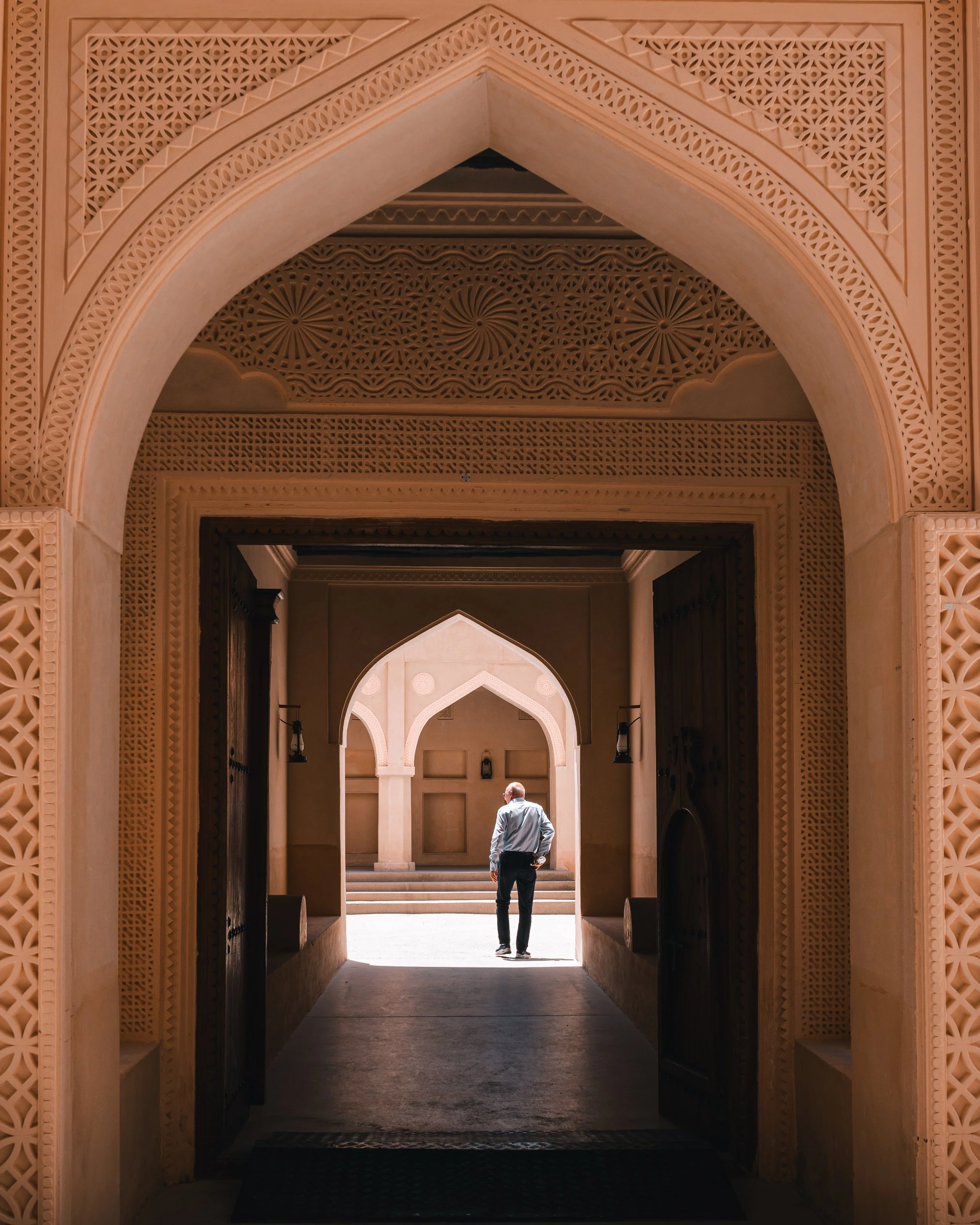 A man in a white shirt and dark pants walking through an ornate arched doorway with intricate architectural details, facing away from the camera.