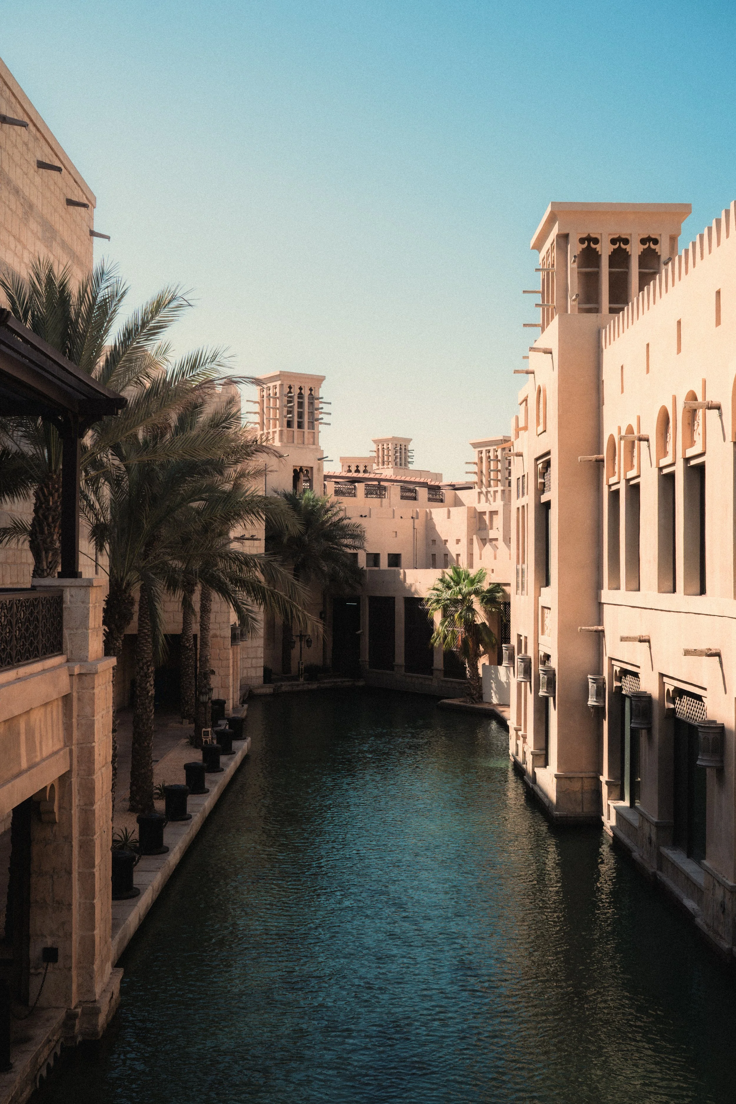 Buildings with Middle Eastern architectural style along a waterway, with palm trees and a clear blue sky.