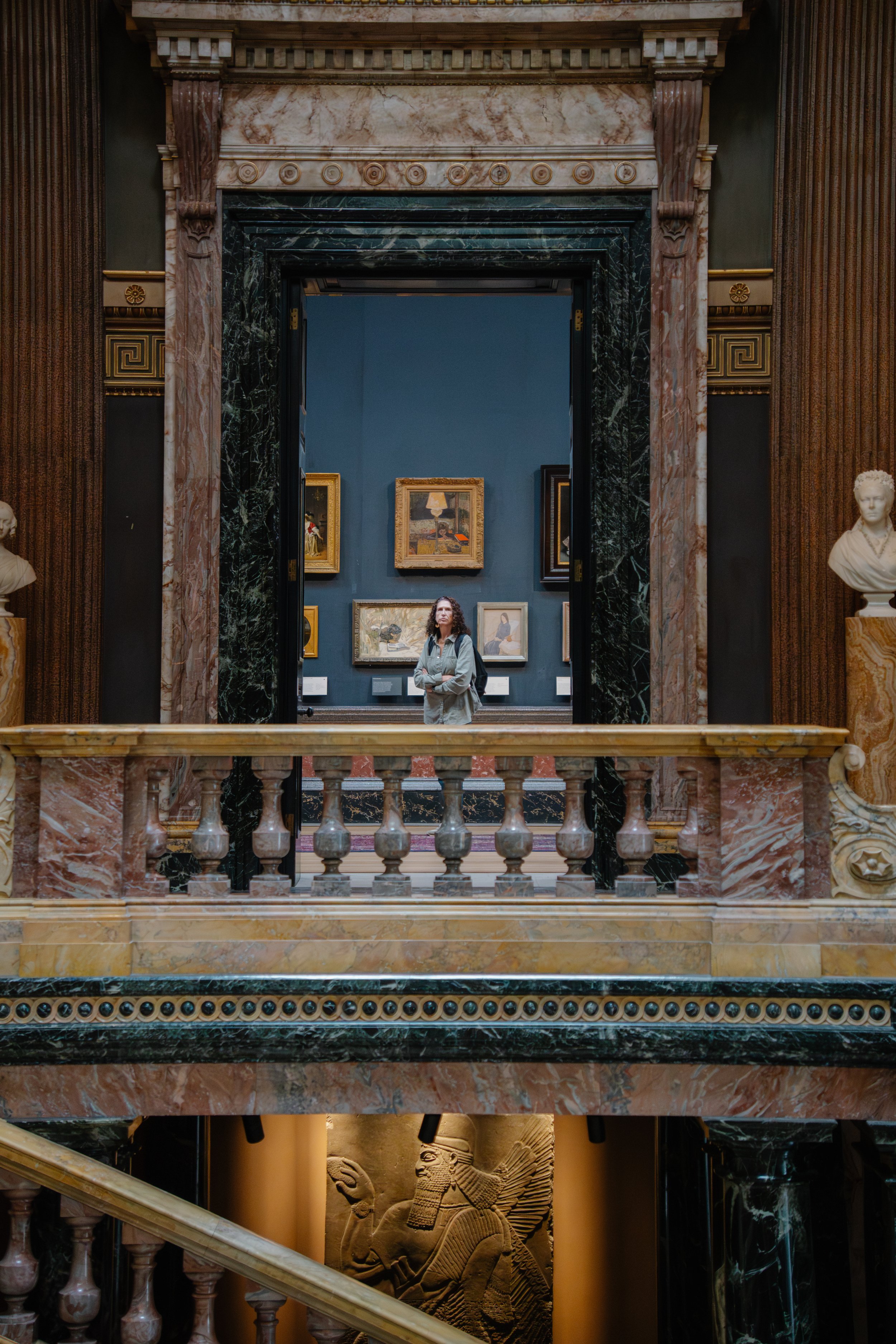 A woman with curly hair and glasses looks at artwork inside a museum hall framed by a building with ornate marble columns and a decorative balustrade.