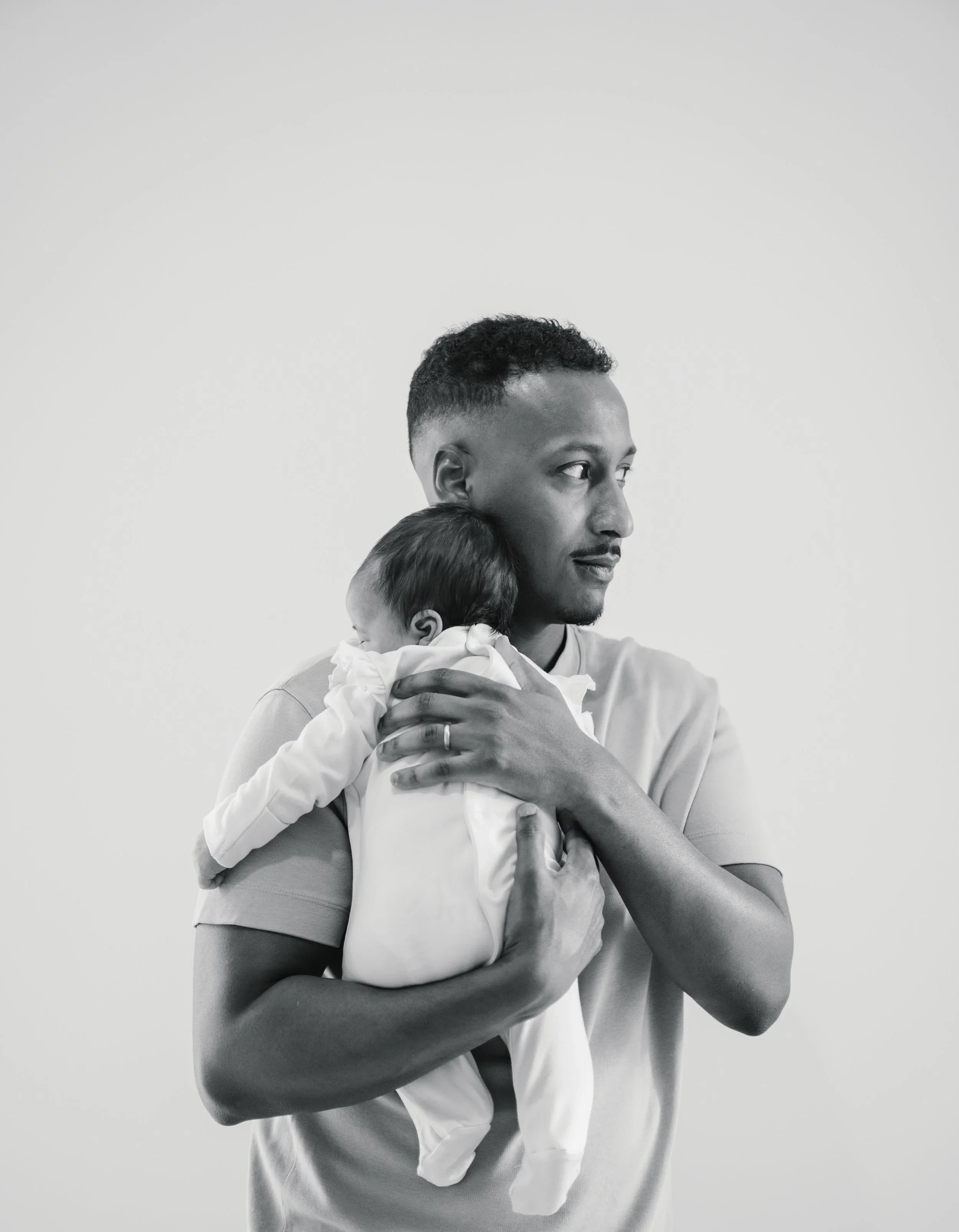 A man holding a sleeping child in his arms, both facing to the right, with a plain white background.