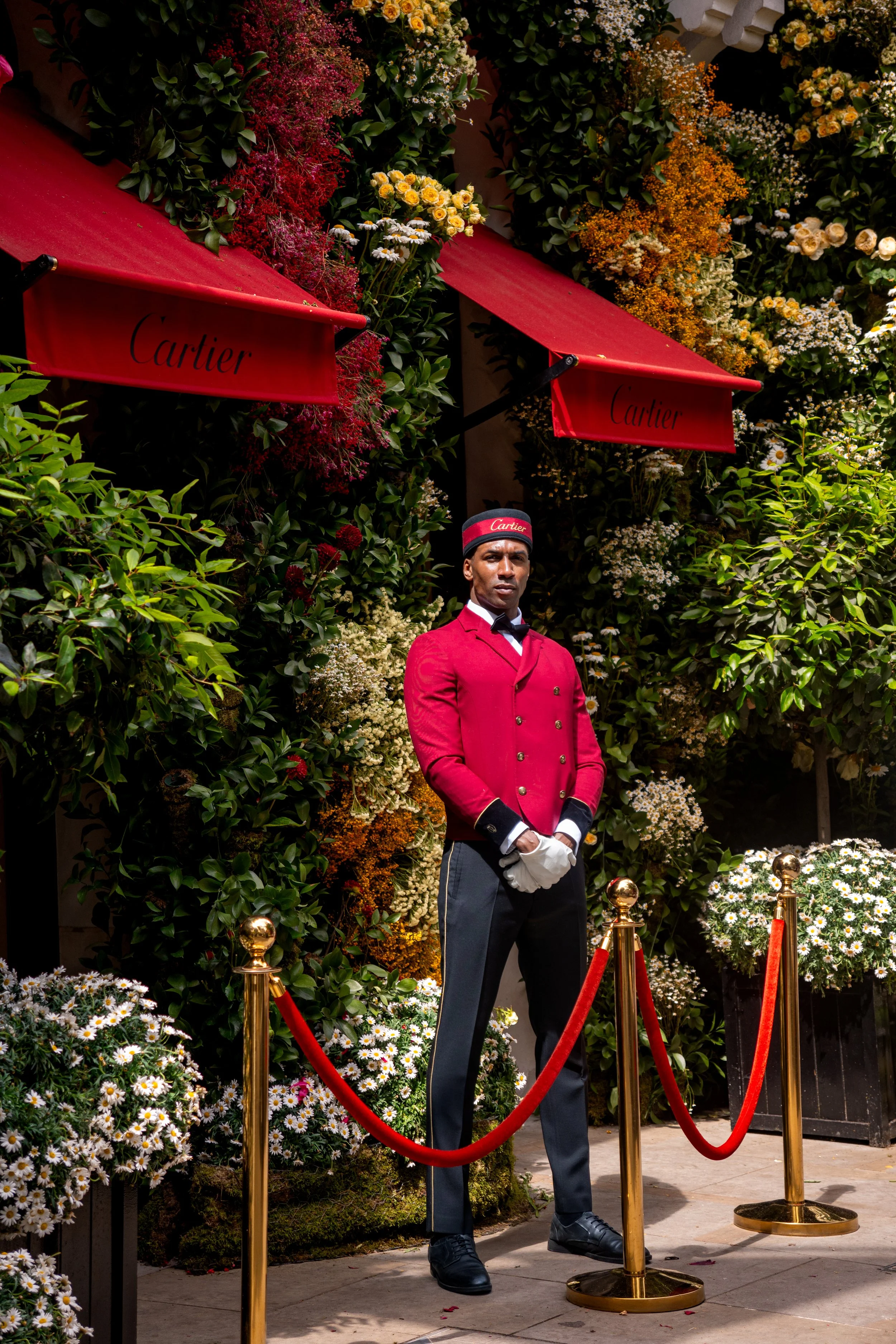 A doorman standing behind a red velvet rope barrier, wearing a red jacket, black gloves, and a black hat with a red band that says 'Cartier'. The background is filled with lush greenery and colorful flowers, with two red awnings labeled 'Cartier' ove