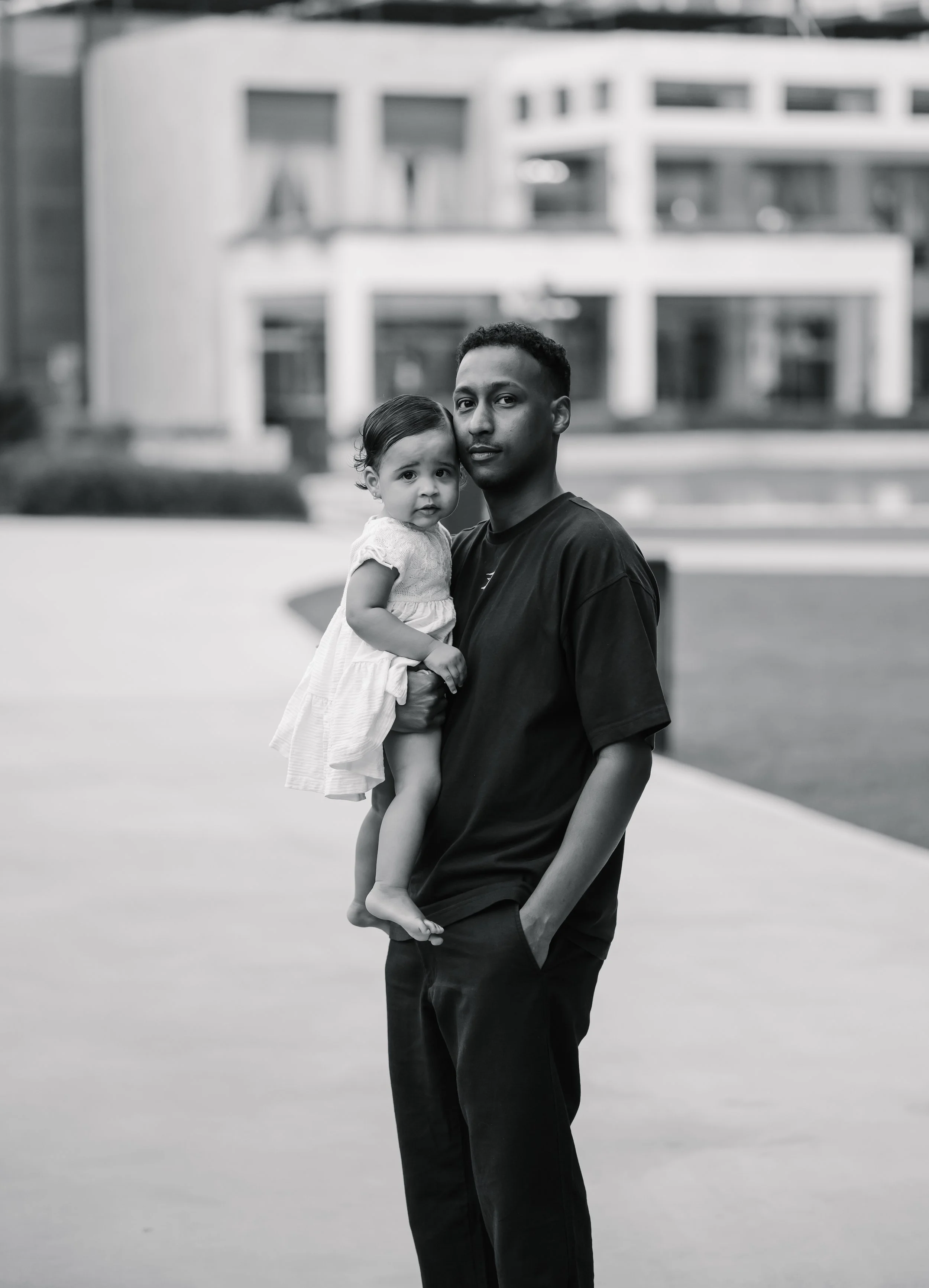 A man holding a young girl in his arms outdoors, in front of a modern building, in black and white.