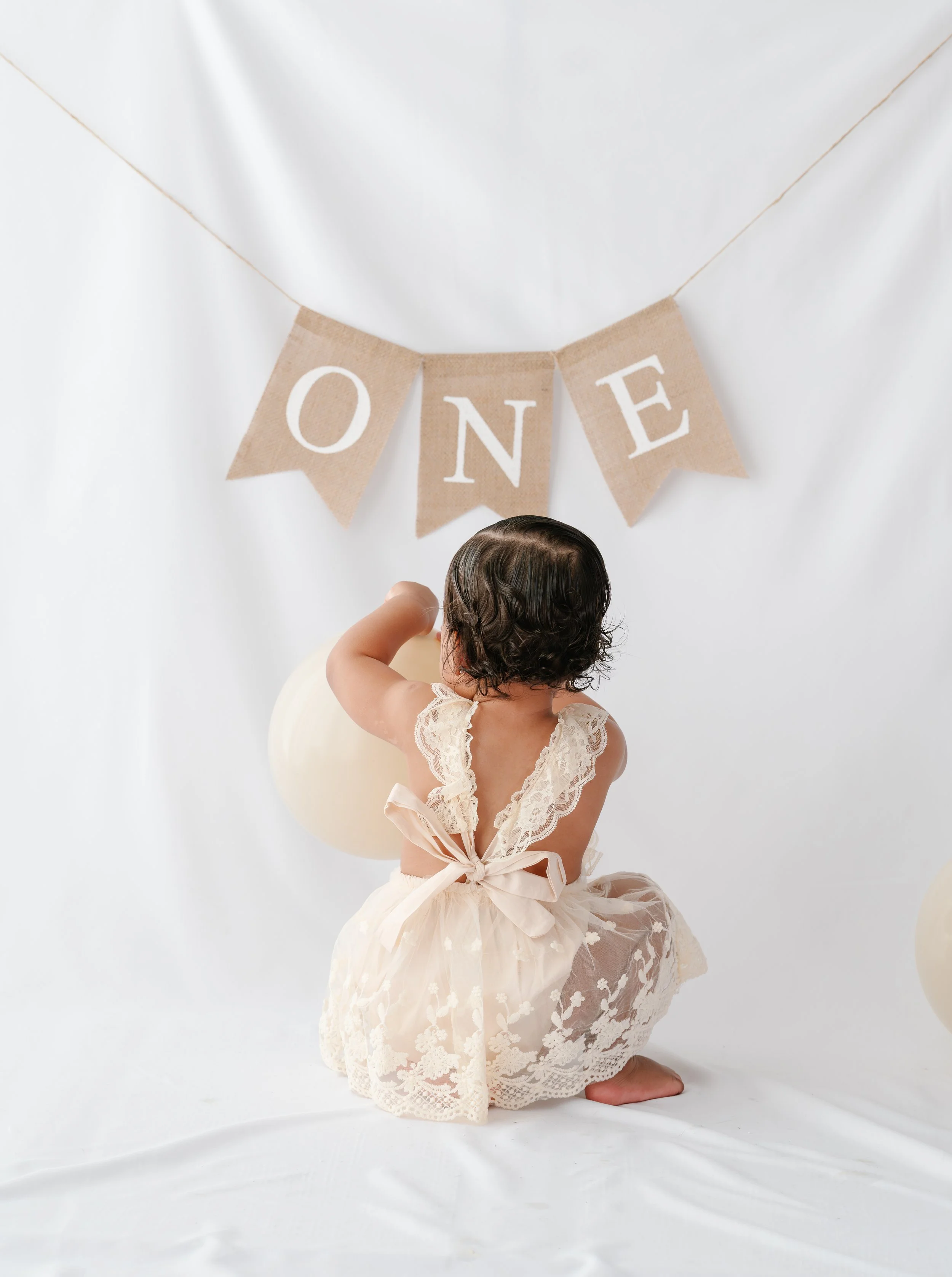 A young girl dressed in cream-colored lace dress with ribbon bow is kneeling on the floor, holding a cream balloon and looking at a beige fabric banner with white letters spelling "ONE" hanging against a white backdrop.