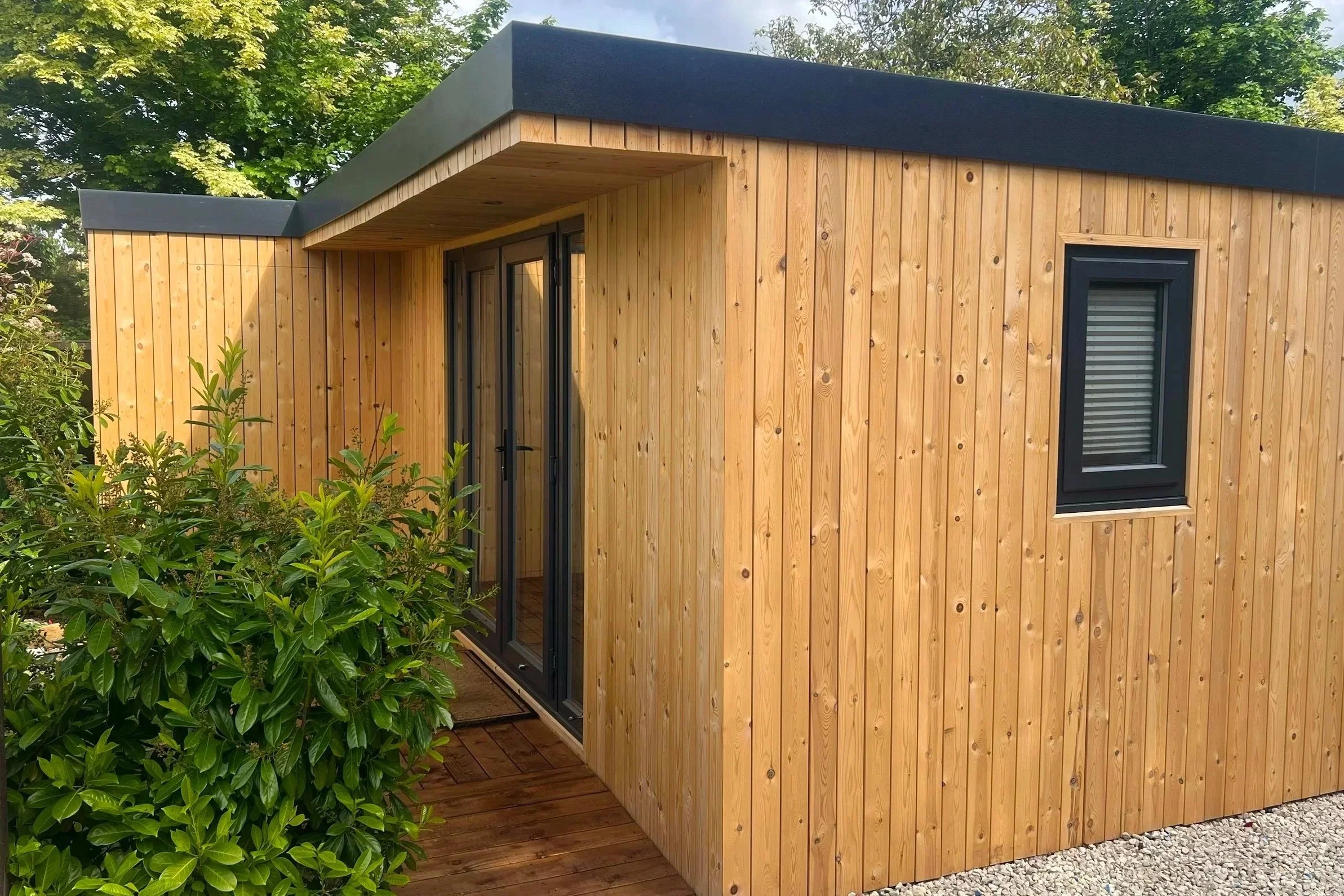 A small wooden exterior structure with vertical wooden planks and black trim around the window and door, surrounded by greenery and plants.
