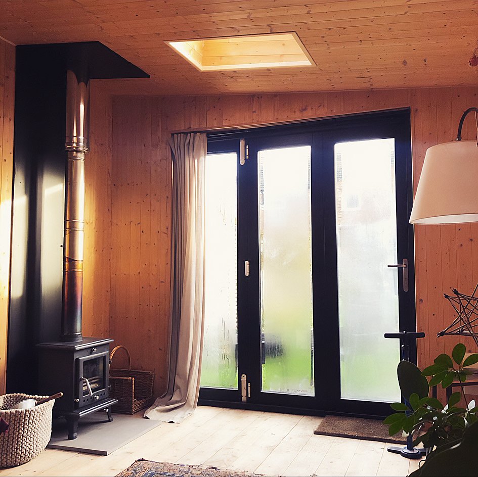Interior of a bespoke garden room looking  cozy room with wooden walls and ceiling, large glass double doors, a wood-burning stove with a chimney pipe, a basket, a curtain, a standing lamp, and a potted plant.