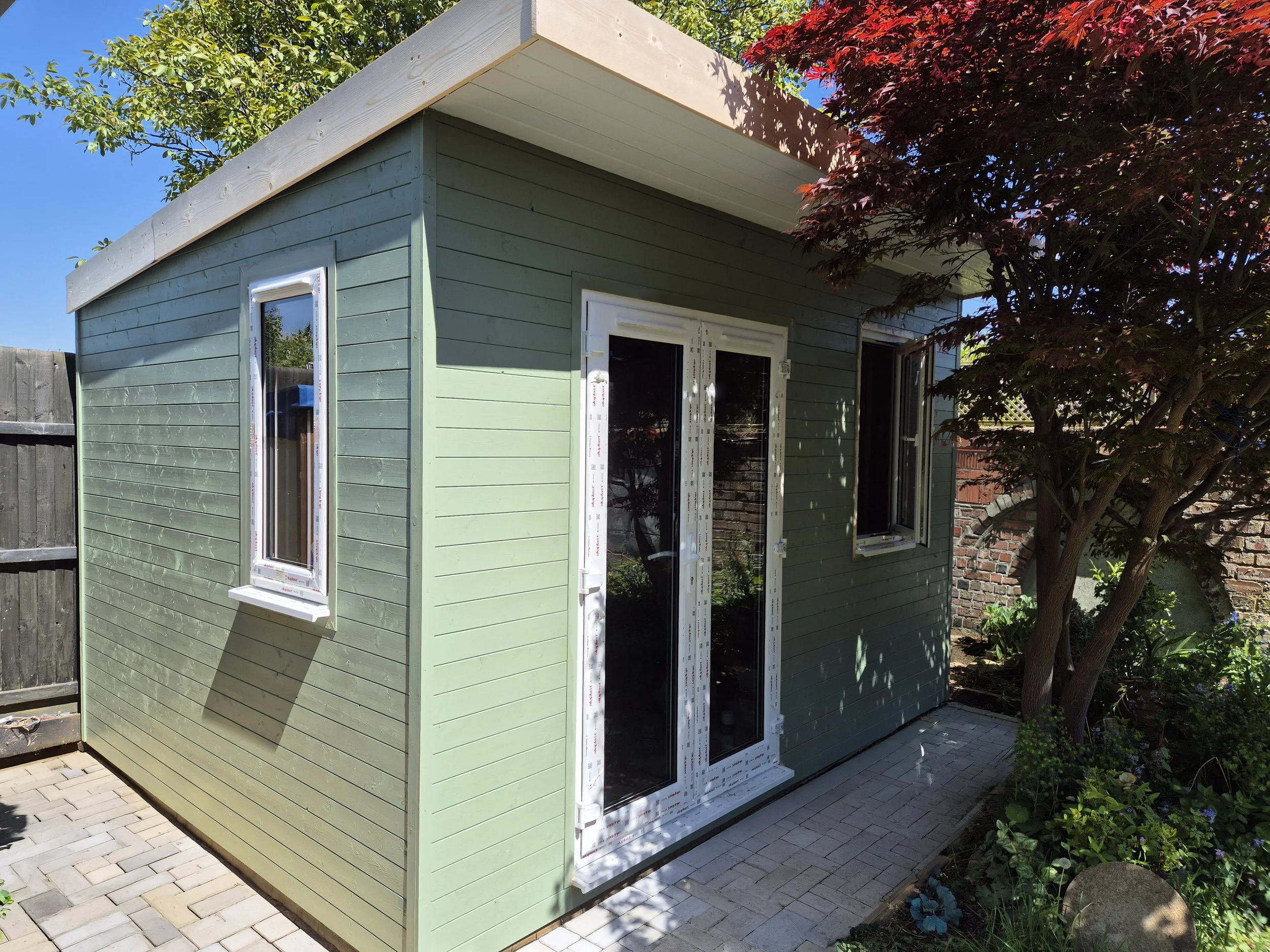 Small bespoke garden room with new windows and siding, surrounded by trees and a paved patio.