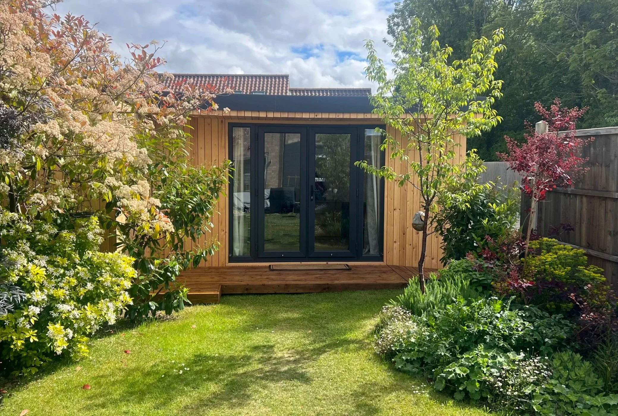 Backyard garden with garden room, surrounded by flowering bushes and trees, under a partly cloudy sky.