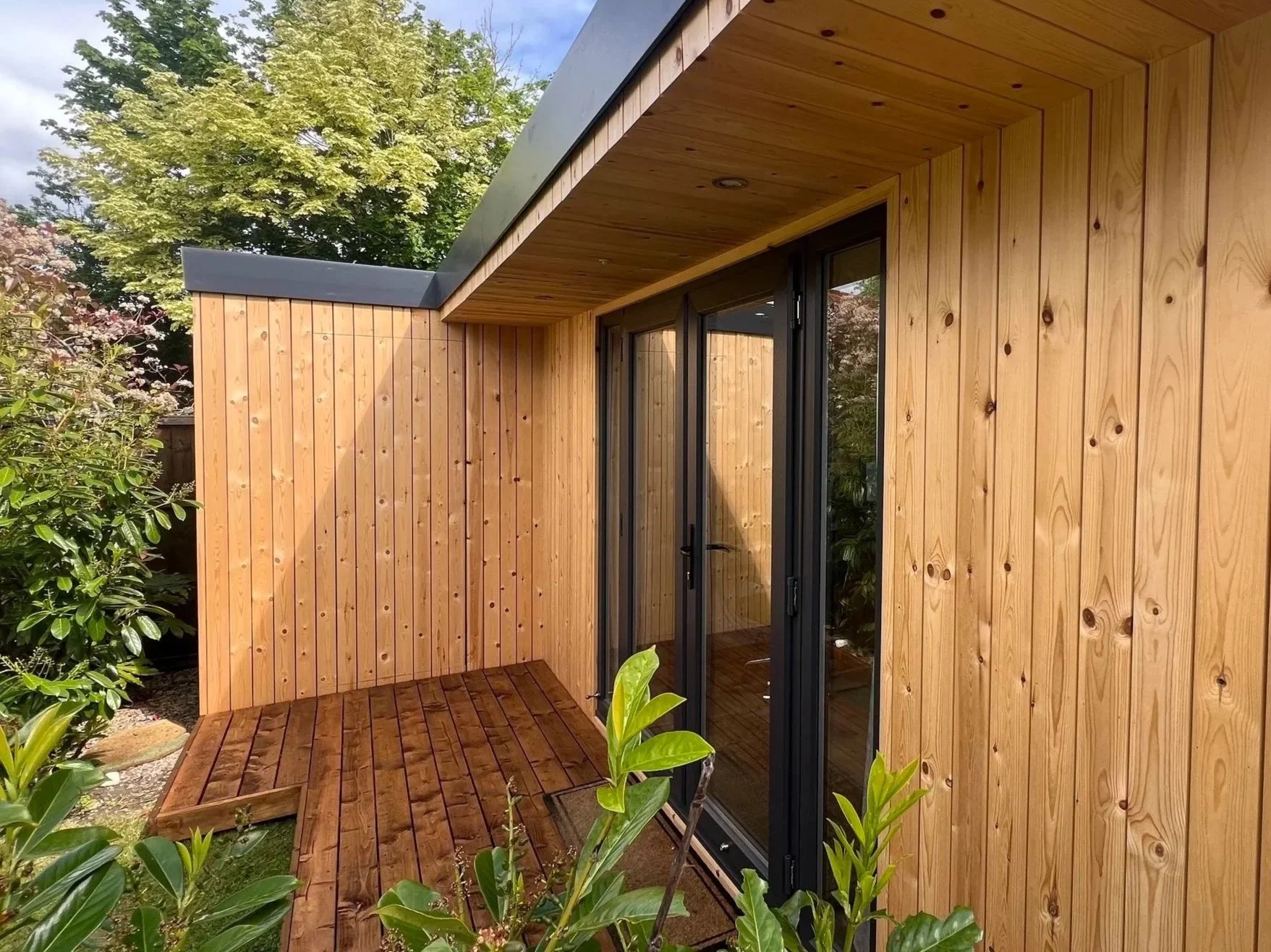 Exterior view of a modern bespoke built garden room with wooden siding, a deck, and a glass door, surrounded by greenery and trees.