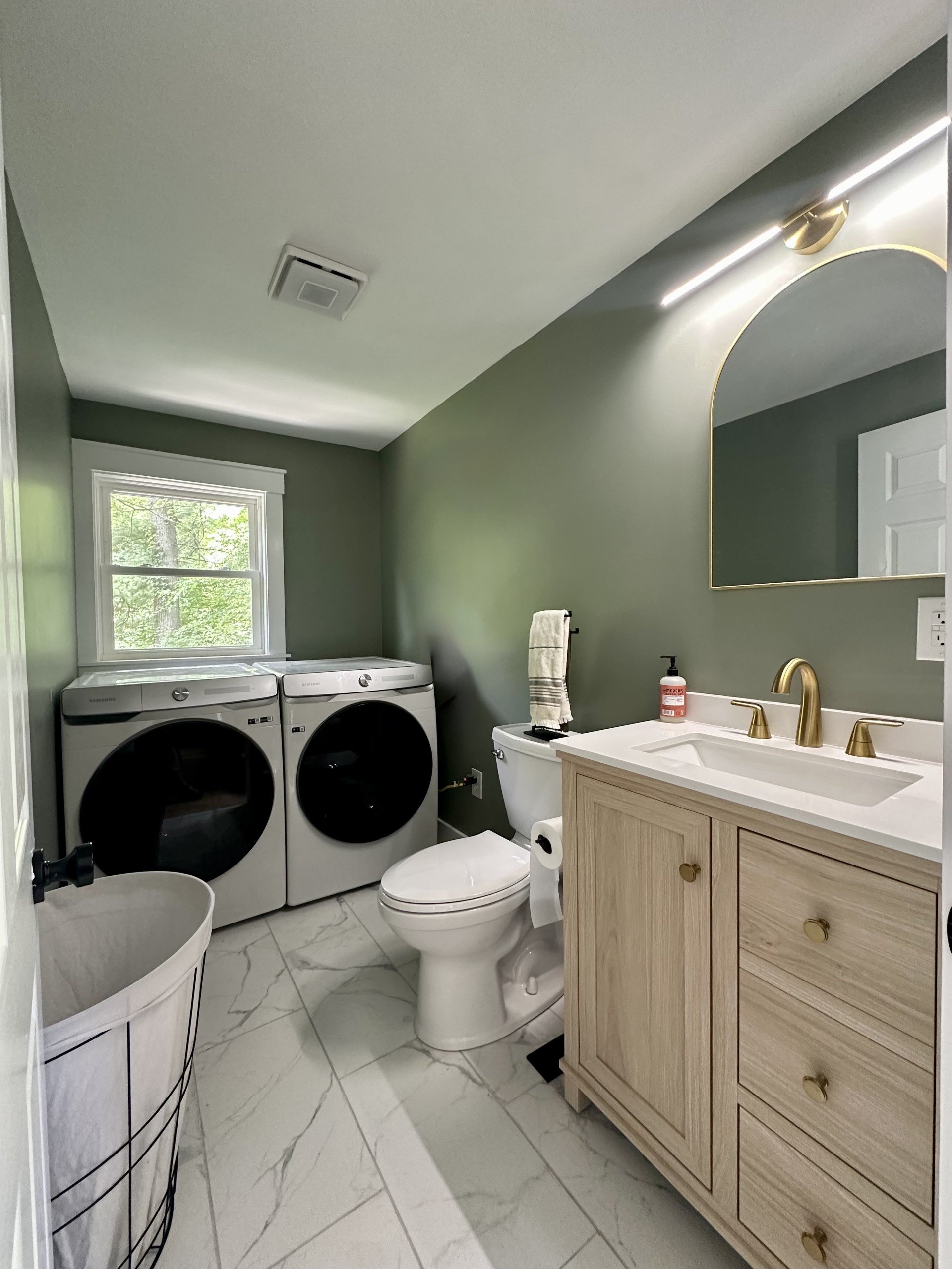A bathroom with a washer and dryer, a toilet, a sink with a gold faucet, a large mirror, and a window with greenery outside.