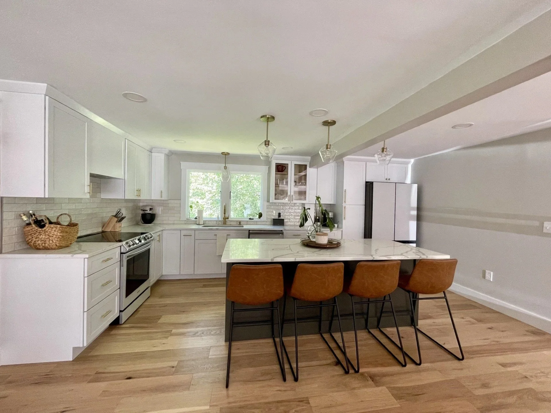 A modern kitchen with white cabinets, a marble island, and a wooden dining table with four brown chairs. Pendant lights hang above the island, and there is a window with greenery outside.