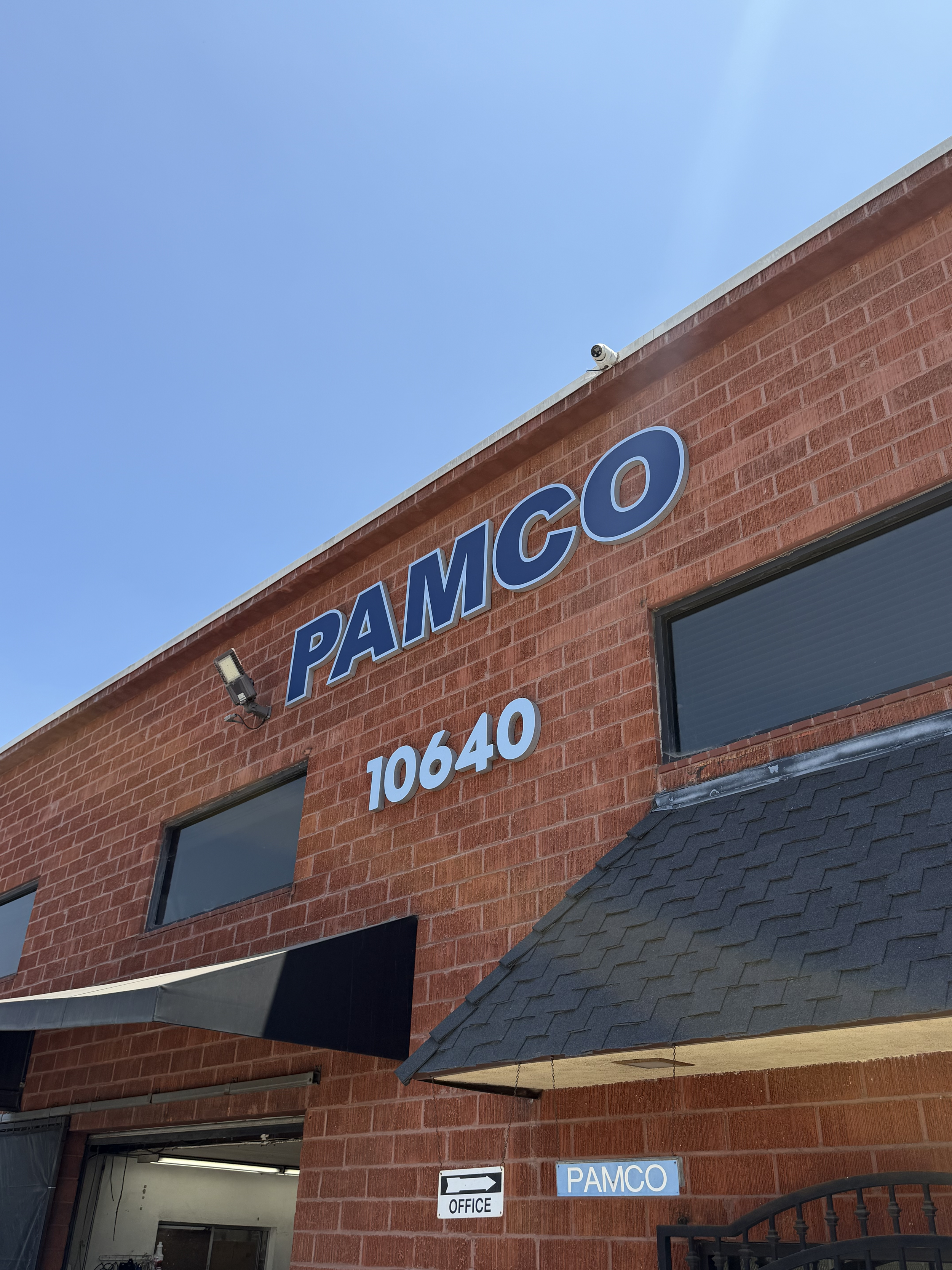 The image shows the exterior of a red brick building with the word 'PAMCO' displayed on the wall in blue letters. There are two rectangular windows, and a black roof overhang with shingles. A small security camera is mounted near the top edge of the building. Below, there is a sign with an arrow labeled 'OFFICE' and the word 'PAMCO' on a blue background.