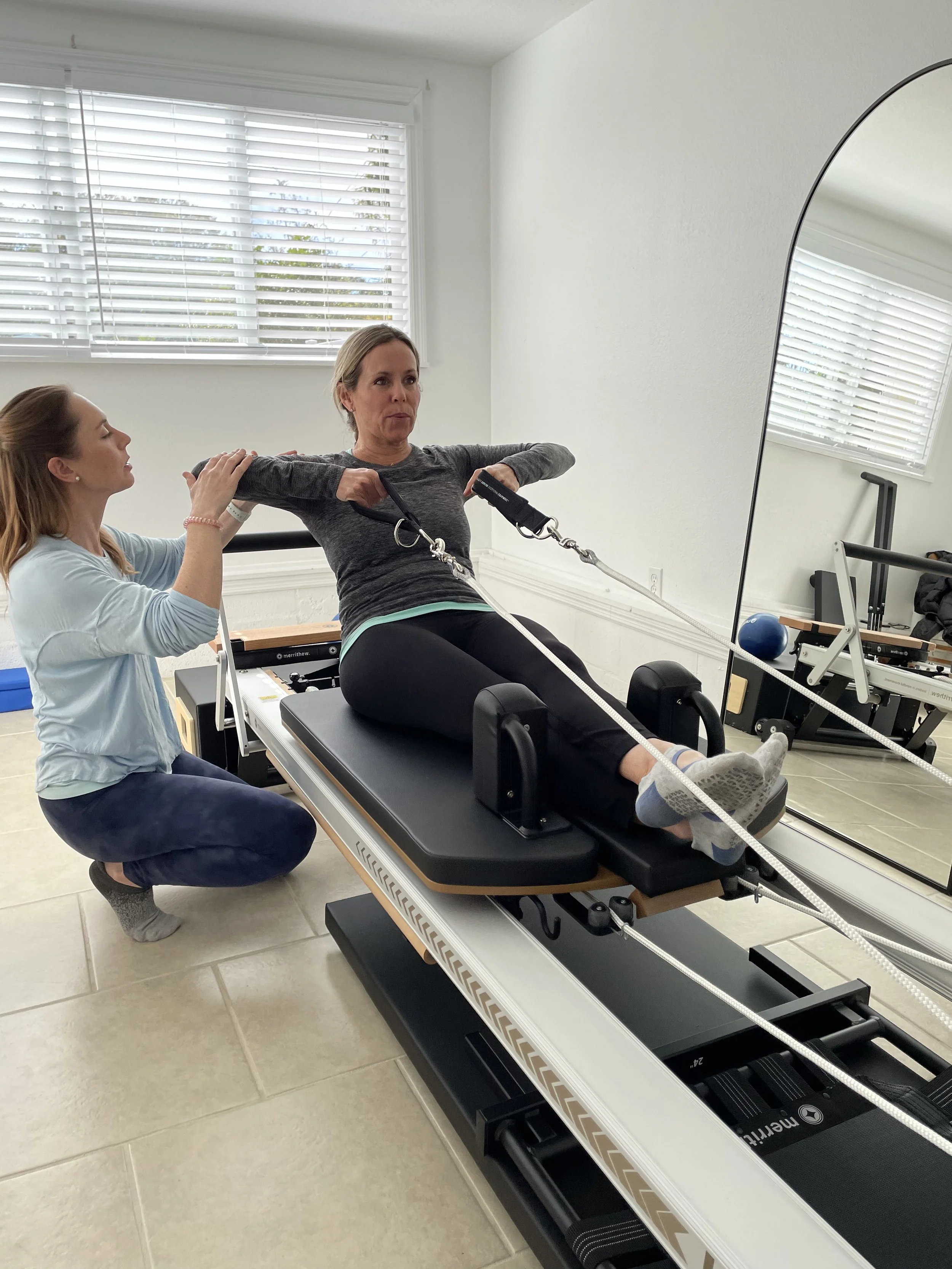 Women doing pilates exercises with Clare, the founder and instructor assisting, using a specialized exercise machine in a bright room with windows.