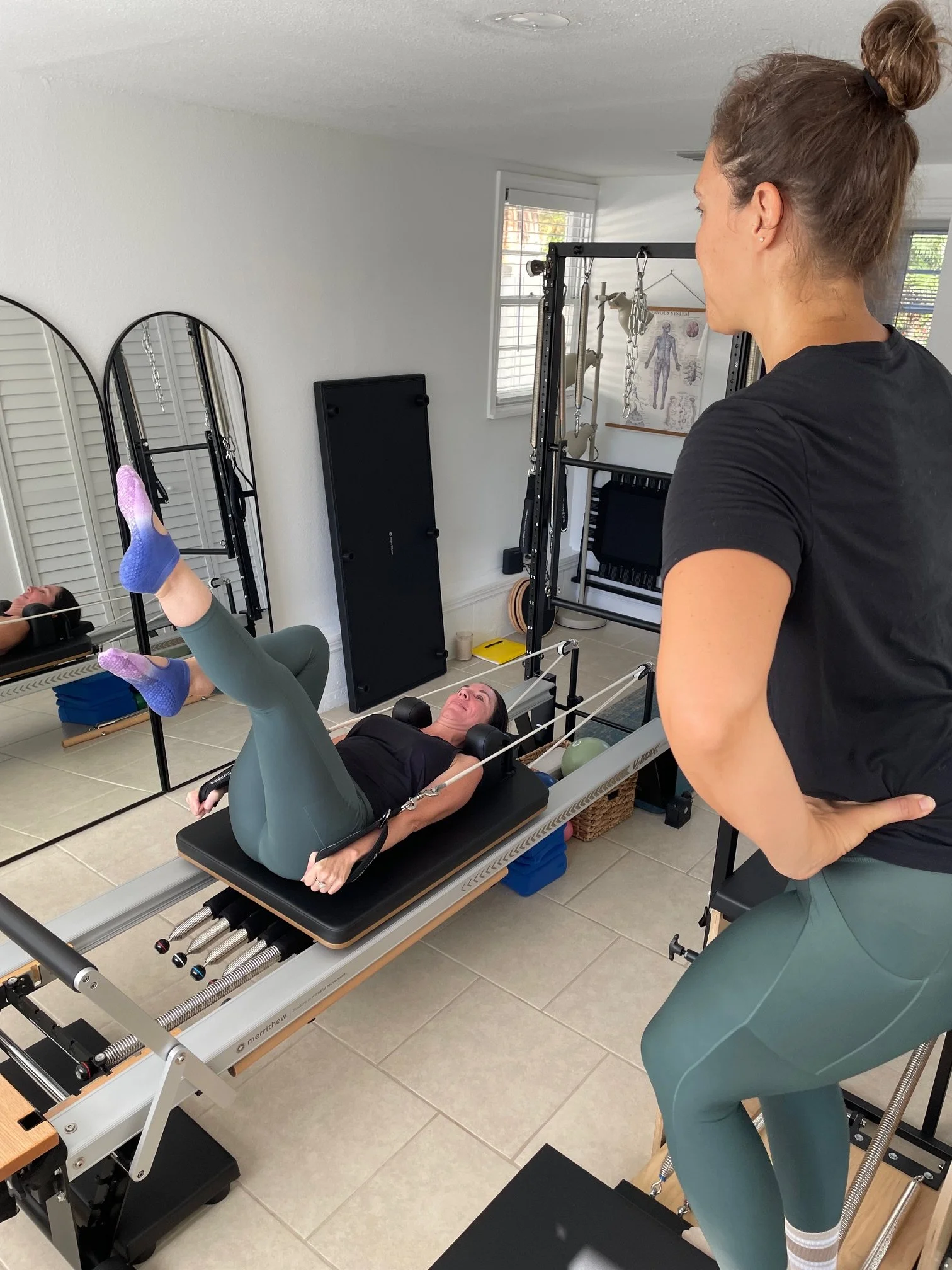 A woman lying on a Pilates reformer machine, with a trainer observing and instructing her during a workout session in a well-lit home gym.