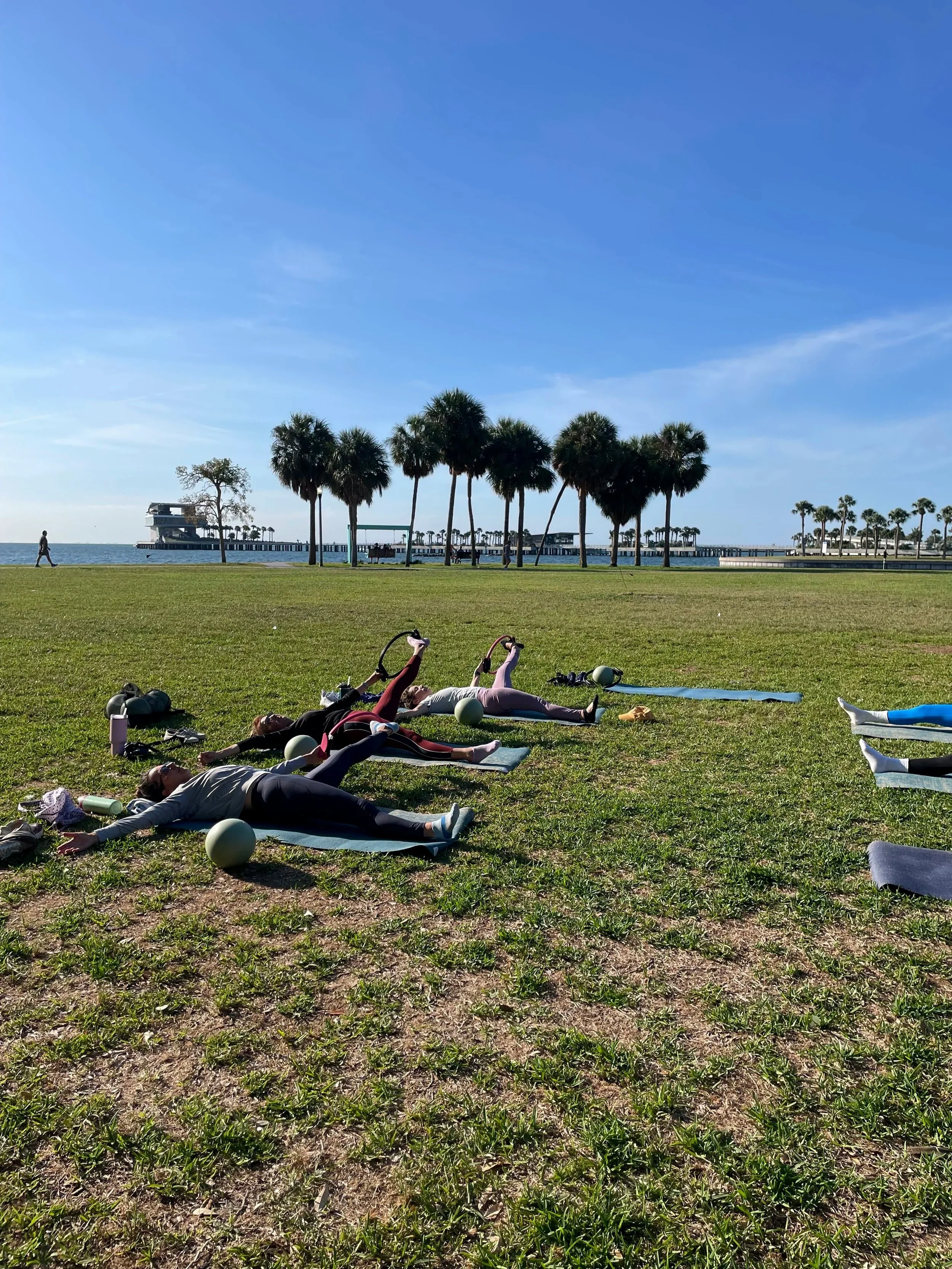 People doing yoga outdoors on a grassy area near the water, with palm trees and a pier in the background under a clear blue sky.