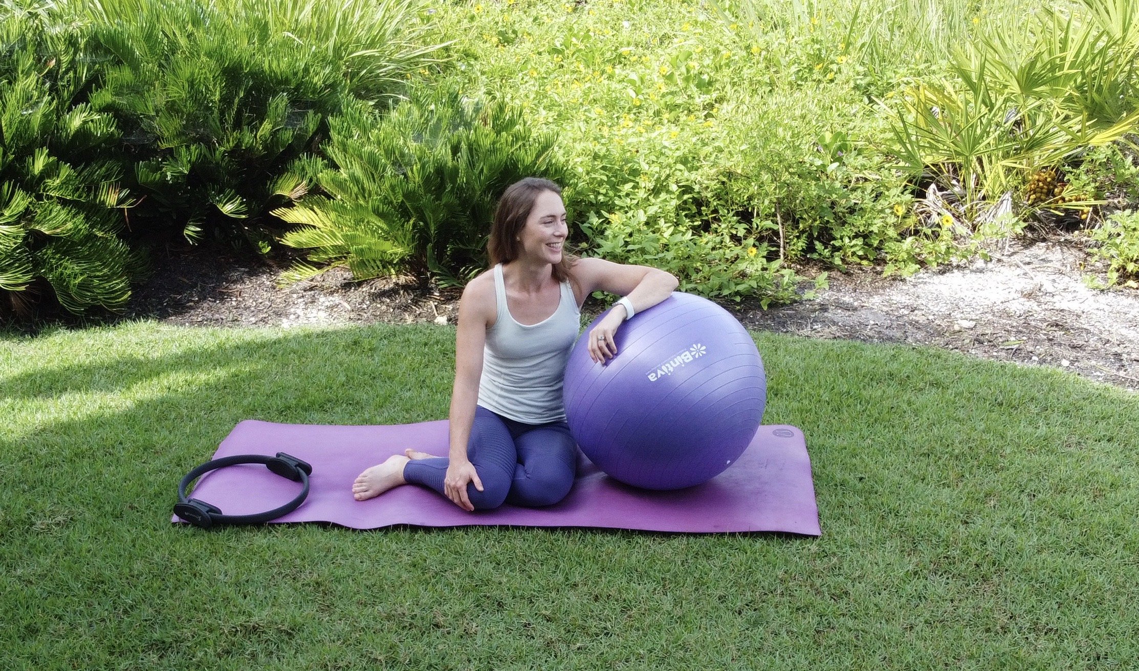 Woman pilates instructor and owner Clare with pilates mat and equipment, on a purple mat, surrounded by greenery,