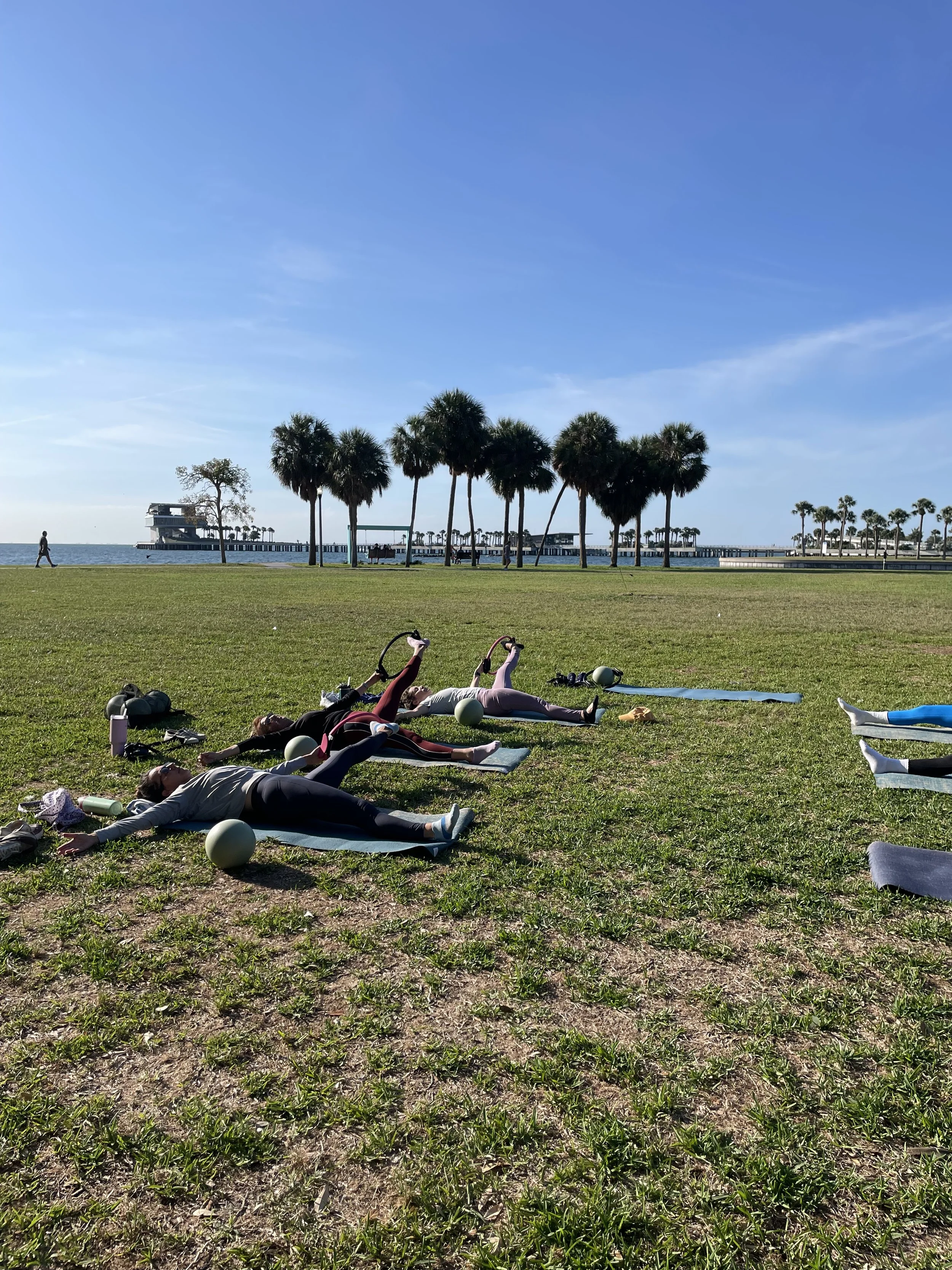 People practicing pilates outdoors in St. Petersburg, FL near the pier on a grassy area with palm trees and a clear blue sky.