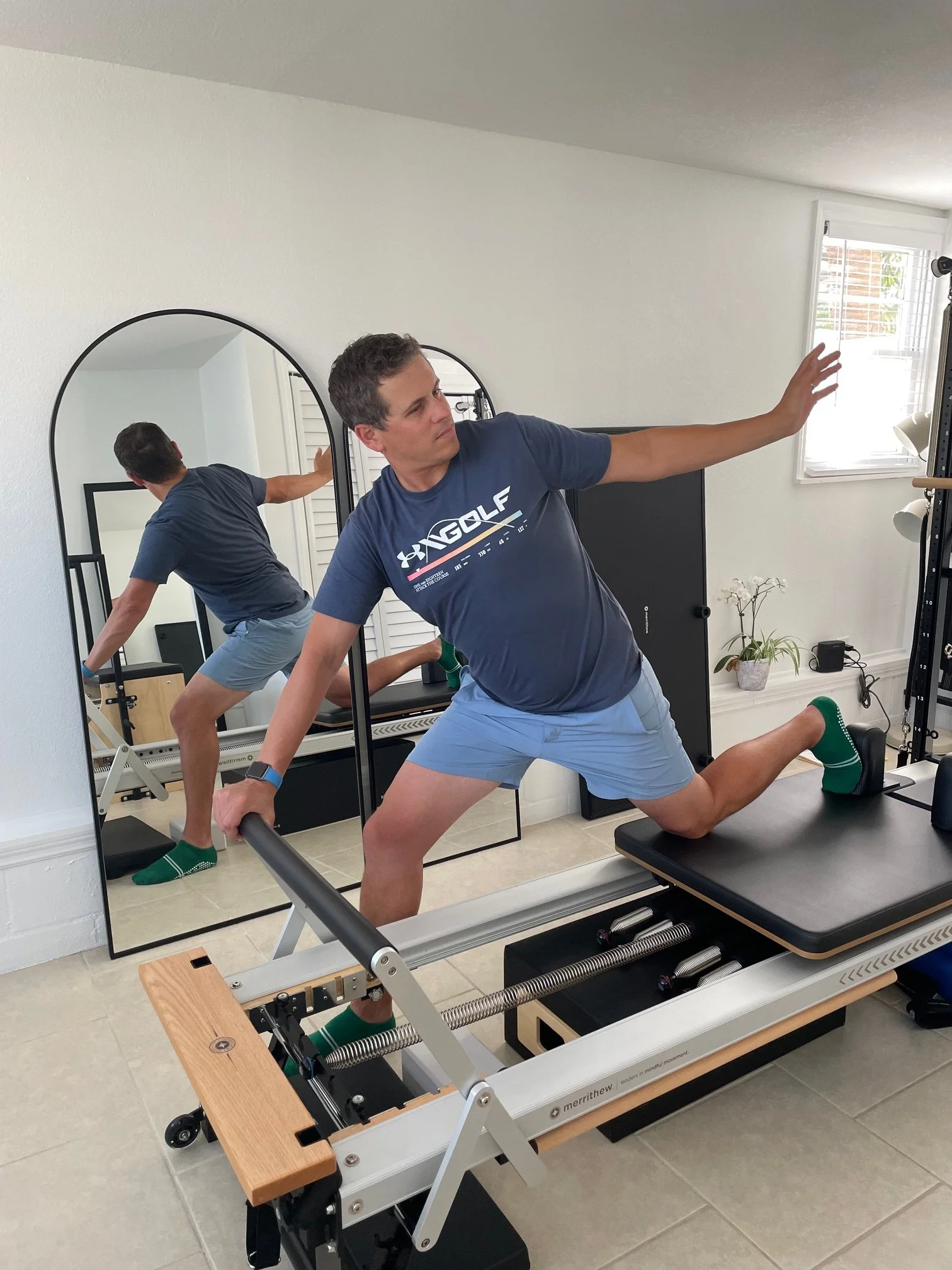 A man stretching in a room with mirrors, fitness equipment, and a window with blinds.