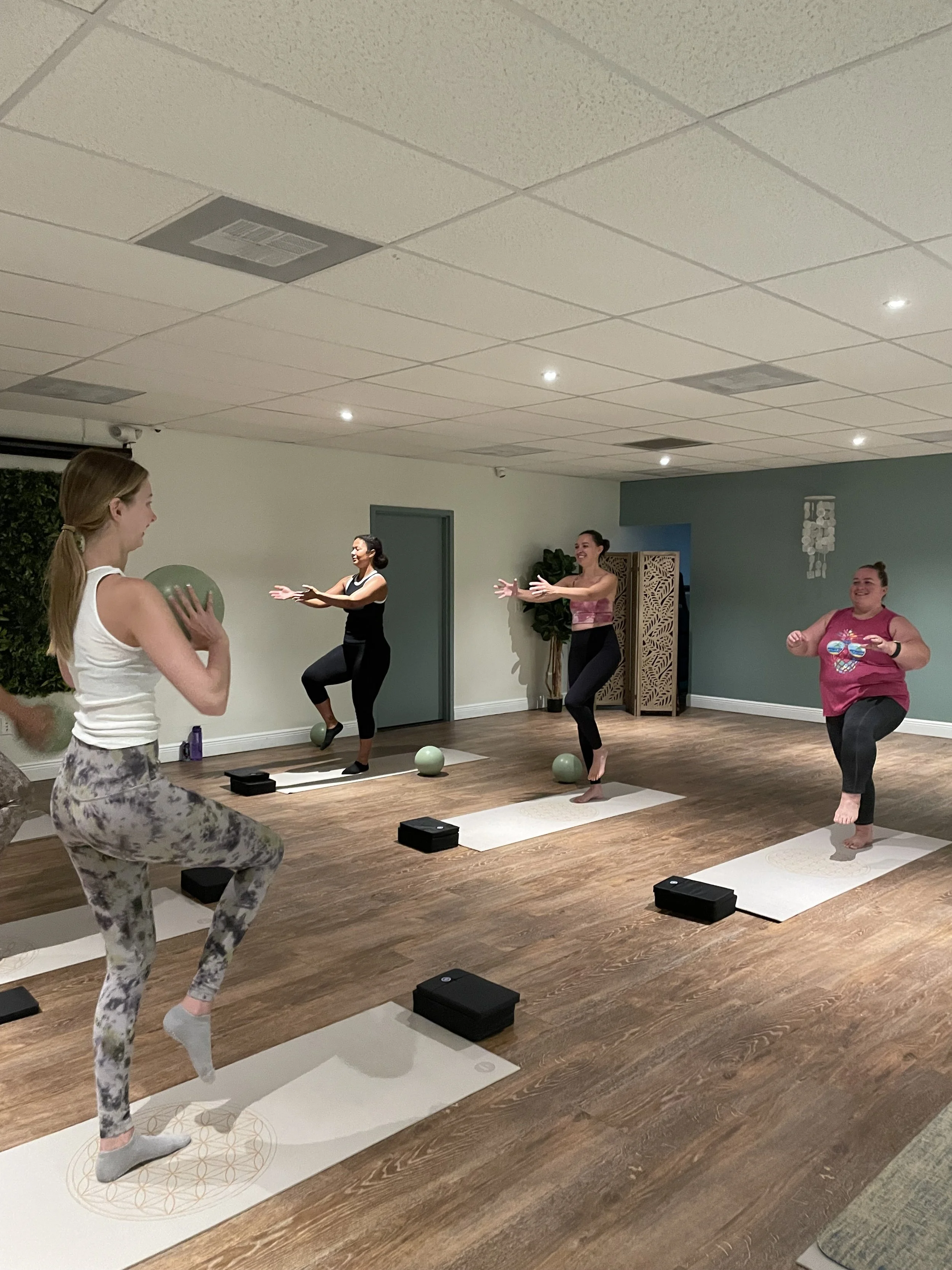 Four women participating in a mat pilates class in a studio with wooden floors, doing balance poses on pilates mats, with exercise balls and blocks nearby.